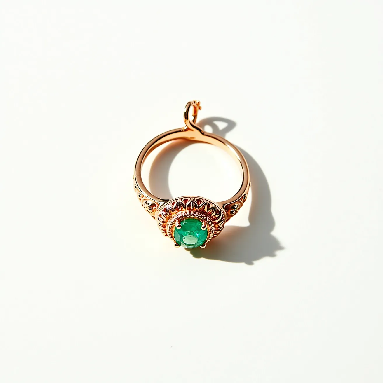 This emerald ring features a vibrant green gemstone, likely an emerald, which is prominently set in the center with an intricate prong setting. The ring is crafted from a metal that appears to be rose gold, providing a warm contrast to the cool green of the emerald. Surrounding the gemstone is a decorative bezel, enhancing the ring's elegance and offering additional security to the central stone. The band is adorned with ornate details, accentuating the overall sophistication of the piece. A small loop or clasp is present, suggesting a versatile design that might allow the ring to be worn as a pendant or attached to another item.