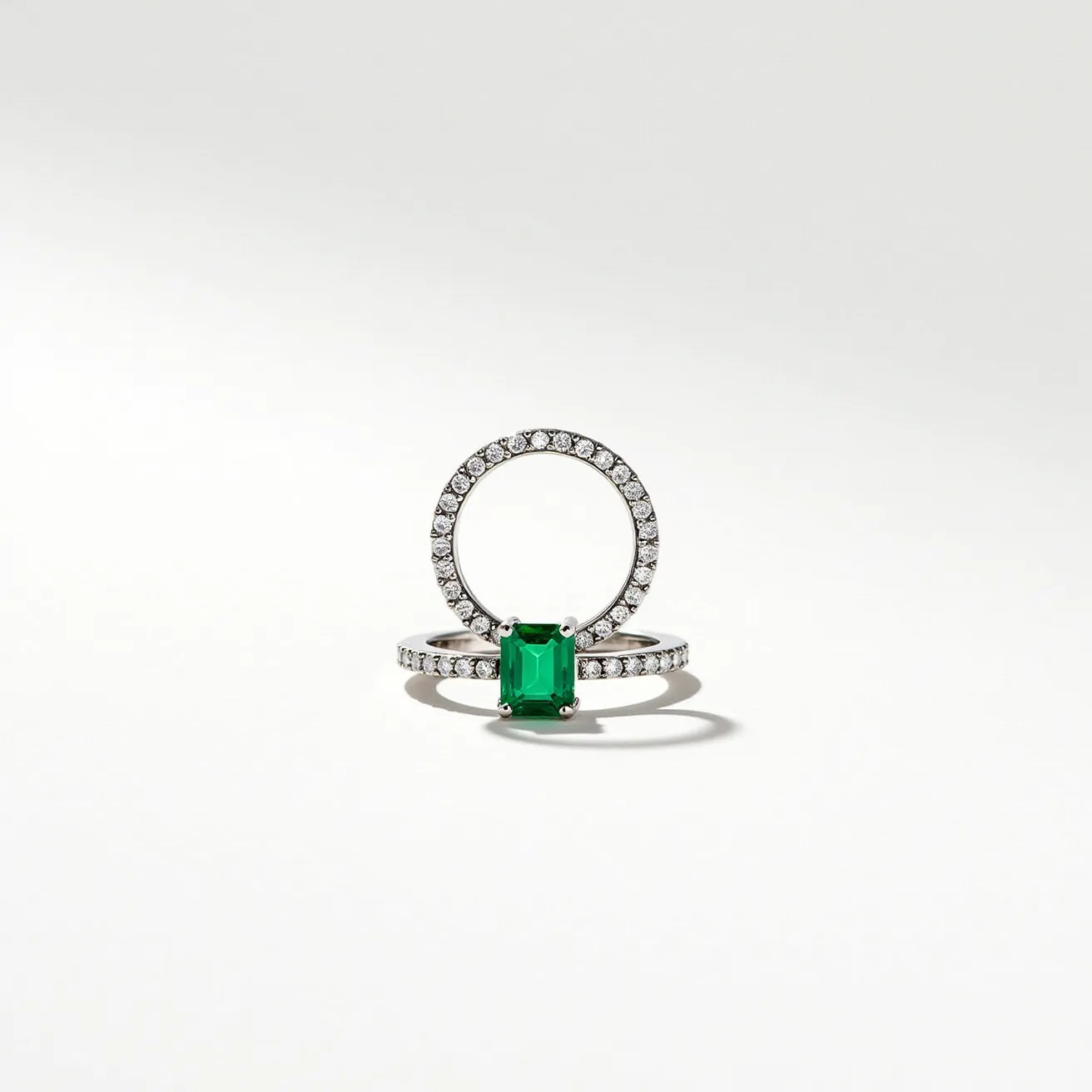 These emerald rings feature a central, striking emerald-cut gem, known for its rectangular shape and stepped facets that accentuate the stone's vibrant green hue. The emerald is set in a prong setting, which securely holds the stone and allows light to pass through, enhancing its brilliance. Surrounding the emerald and adorning the band are multiple small round diamonds, each in a pavé setting that creates a continuous sparkle along the metal. The rings are crafted from a lustrous metal, likely platinum or white gold, which complements the vivid green of the emerald and the sparkle of the diamonds. The combination of elements adds a classic yet luxurious touch to the overall design.