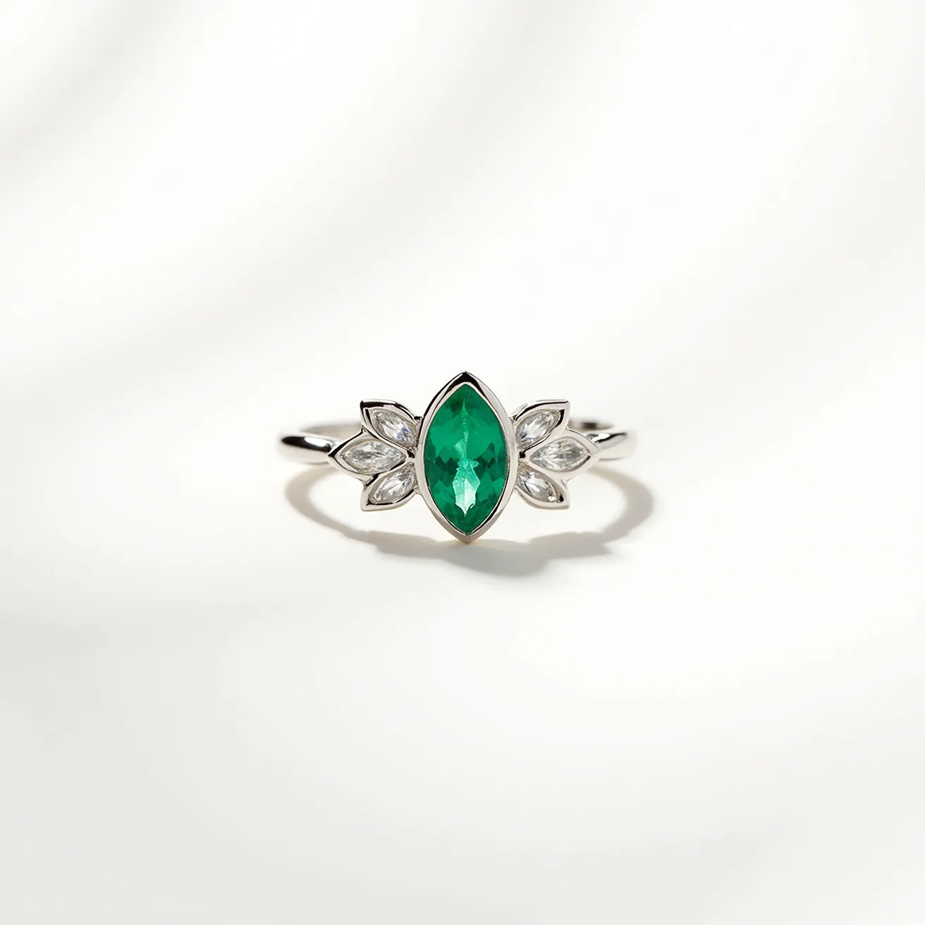 This emerald ring features a captivating marquise-cut emerald as the central gemstone, set in a secure bezel-style setting. Flanking the emerald are small marquise-cut white gemstones, likely diamonds, arranged in a leaf-like or petal design, enhancing the ring's elegance and symmetry. The band appears to be crafted from a polished metal, likely white gold or platinum, adding a sleek and sophisticated finish to the ring. The combination of the vibrant green emerald and the sparkling white gemstones creates a striking and luxurious piece of jewelry.