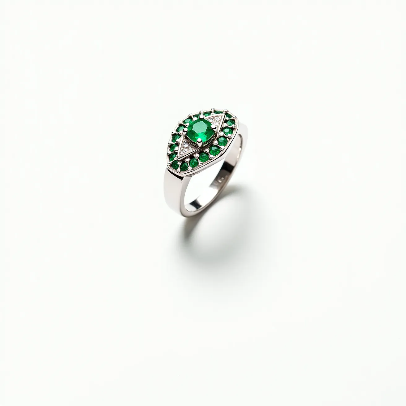 This emerald ring features a central oval-cut emerald, surrounded by a symmetrical halo of smaller round emeralds, all set in a sleek metal band, likely platinum or white gold. The emeralds are securely held in a prong setting, enhancing the stones' visibility and brilliance. The ring showcases an intricate design with a triangular accent complementing the central emerald, contributing to its luxurious and elegant appearance.