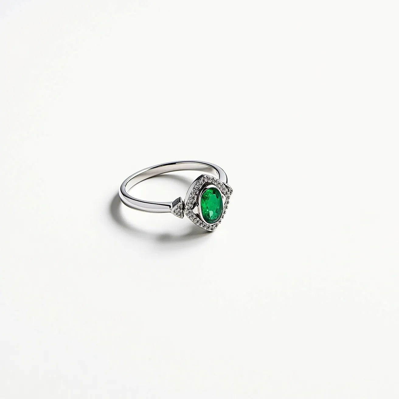 This emerald ring features a central, oval-cut emerald set in a delicate band. The emerald is surrounded by smaller clear stones, likely diamonds, arranged in a halo setting that accentuates the rich green of the central gem. The ring's band appears to be crafted from a silvery metal, possibly white gold or platinum, offering a sleek and elegant look. The setting complements the ring’s overall design, providing a refined and classic aesthetic ideal for various occasions.