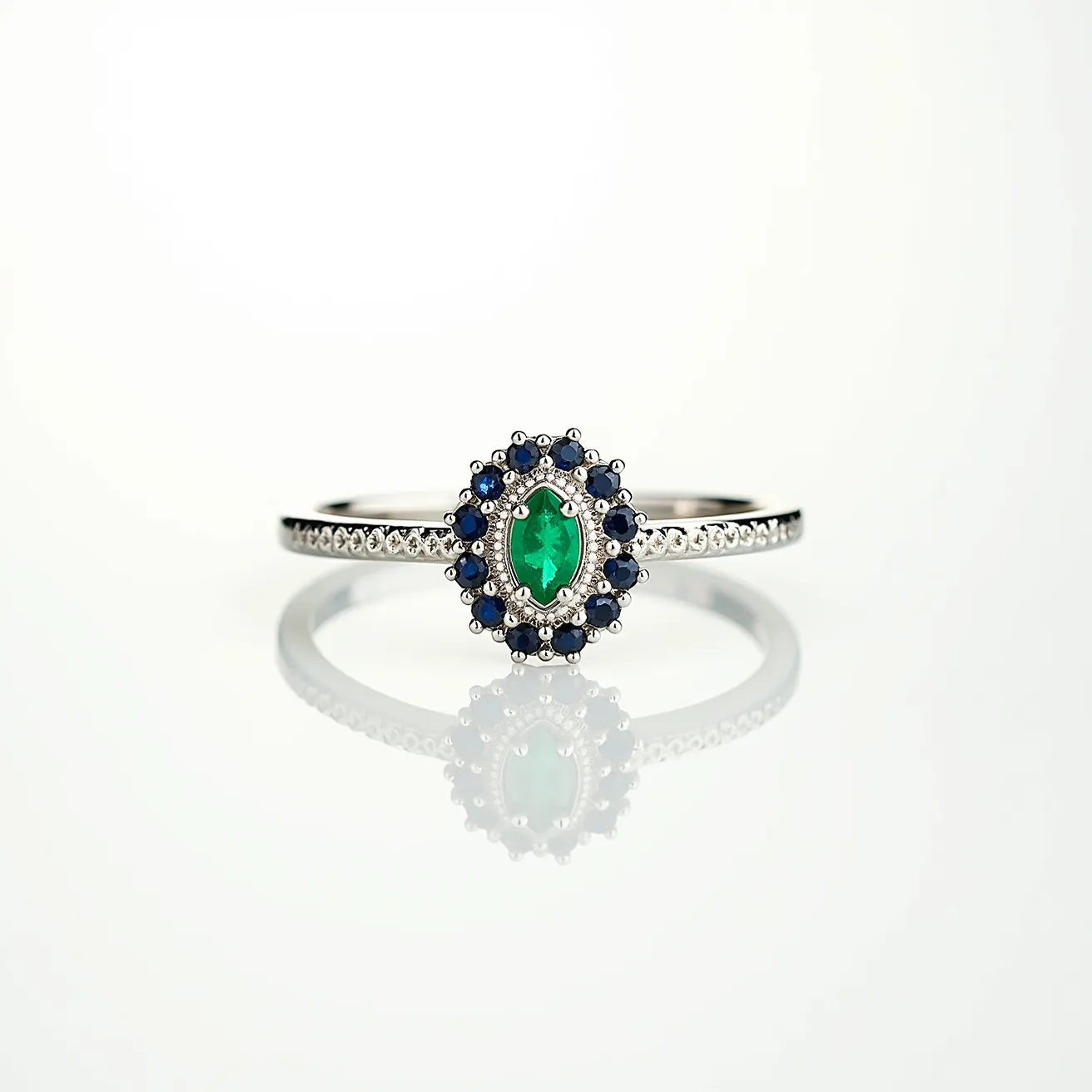 This emerald ring showcases a striking oval-cut emerald at its center, surrounded by a halo of smaller round-cut blue sapphires. The stones are set on a sleek band, likely made of white gold or platinum, which adds to its elegance. The band is adorned with a row of small diamonds, enhancing the ring’s overall brilliance and luxury. The setting is classic, highlighting the vibrancy of the emerald while the sapphires provide a beautiful contrast.