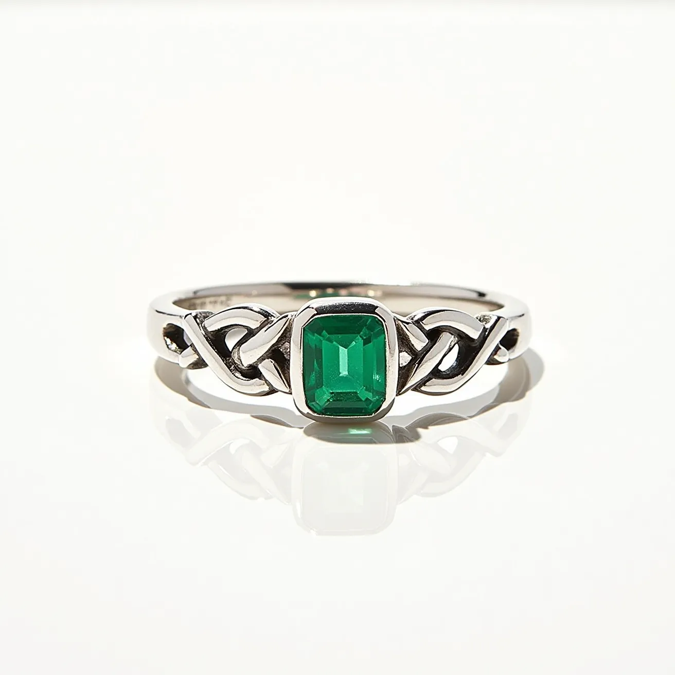 This emerald ring features a striking rectangular emerald-cut gemstone set prominently in the center, showcasing a deep green hue characteristic of high-quality emeralds. The setting appears to be crafted from a polished metal, likely white gold or platinum, which enhances the vibrant color of the emerald. The band exhibits an intricate Celtic-inspired design with intertwining knots, adding an elegant and symbolic touch to the piece. The ring's craftsmanship highlights both the beauty of the gemstone and the skillful detailing of the metalwork, making it a unique and captivating accessory.