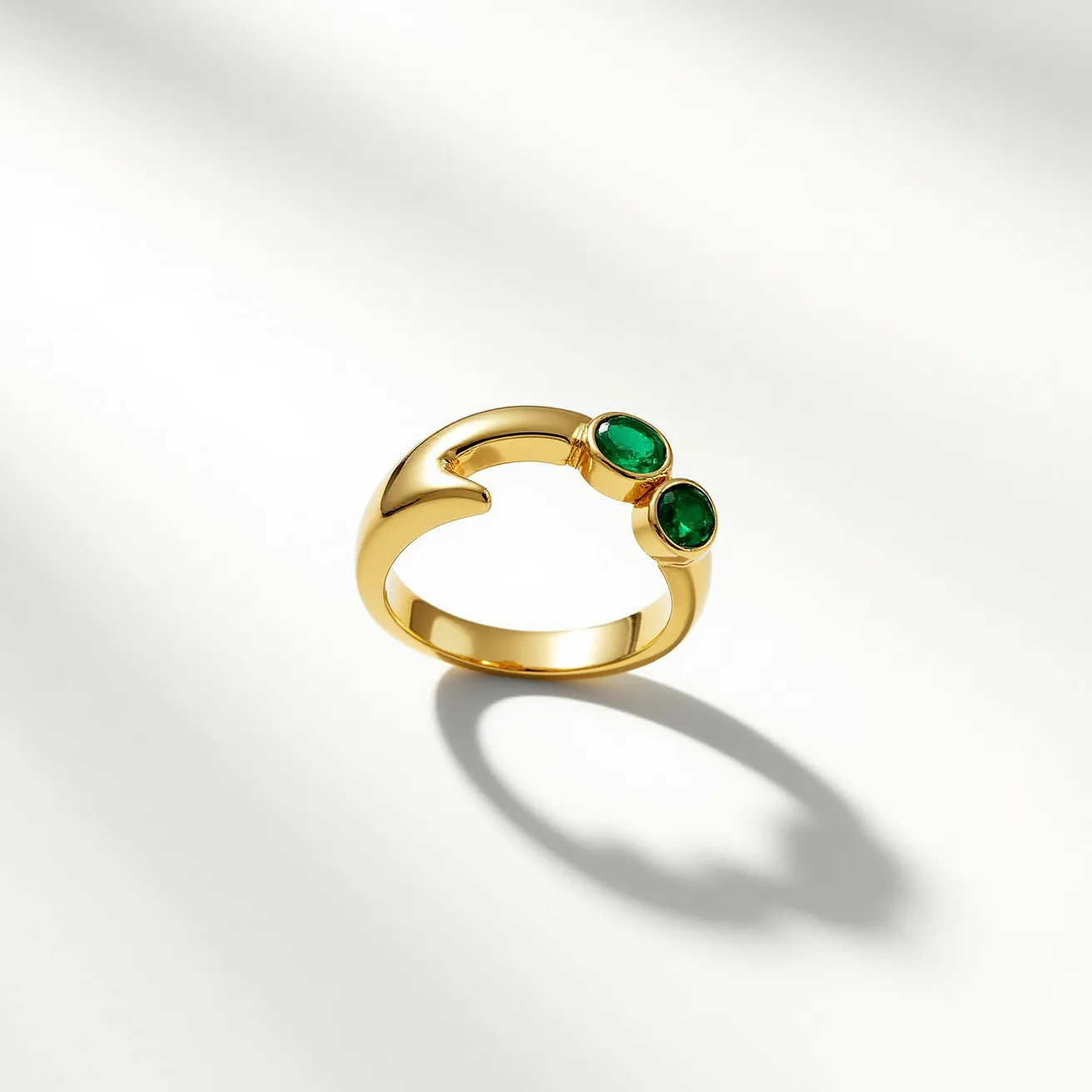 This emerald ring features a sleek, polished gold band that gracefully curves into an open-ended design. It elegantly showcases two vibrant green emeralds, each with an oval cut. The emeralds are set securely in bezel settings, allowing the stones to be prominently displayed while providing a protective border. The ring's open-ended structure adds a modern touch, enhancing the overall aesthetic appeal of the piece.