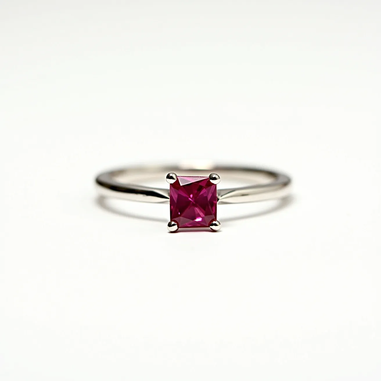 This engagement ring features a sleek metal band, likely made of white gold or platinum, and showcases a vibrant square-cut pink gemstone, possibly a ruby or pink sapphire, held securely in a four-prong setting. The prongs are crafted to elegantly secure the stone while maximizing its exposure to light, enhancing its brilliance. The band is simple and slender, emphasizing the central stone as the focal point of the design.