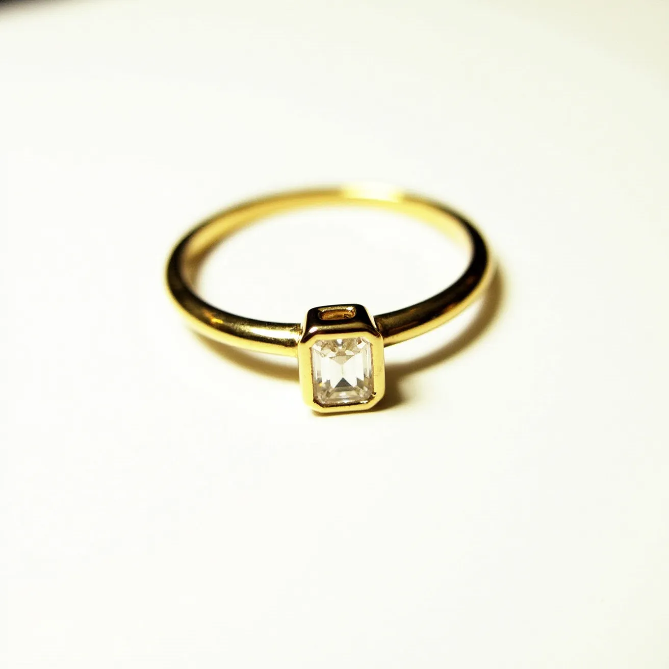 This engagement ring features a sleek, minimalist design with a yellow gold band. The centerpiece is a clear, emerald-cut gemstone, held securely in a bezel setting, which provides a streamlined and modern aesthetic. The band appears to be polished, enhancing the brilliance of the gold, and complementing the angular facets of the stone. The combination of the gold band and the geometric precision of the emerald-cut gem creates a sophisticated and timeless look suitable for any engagement.