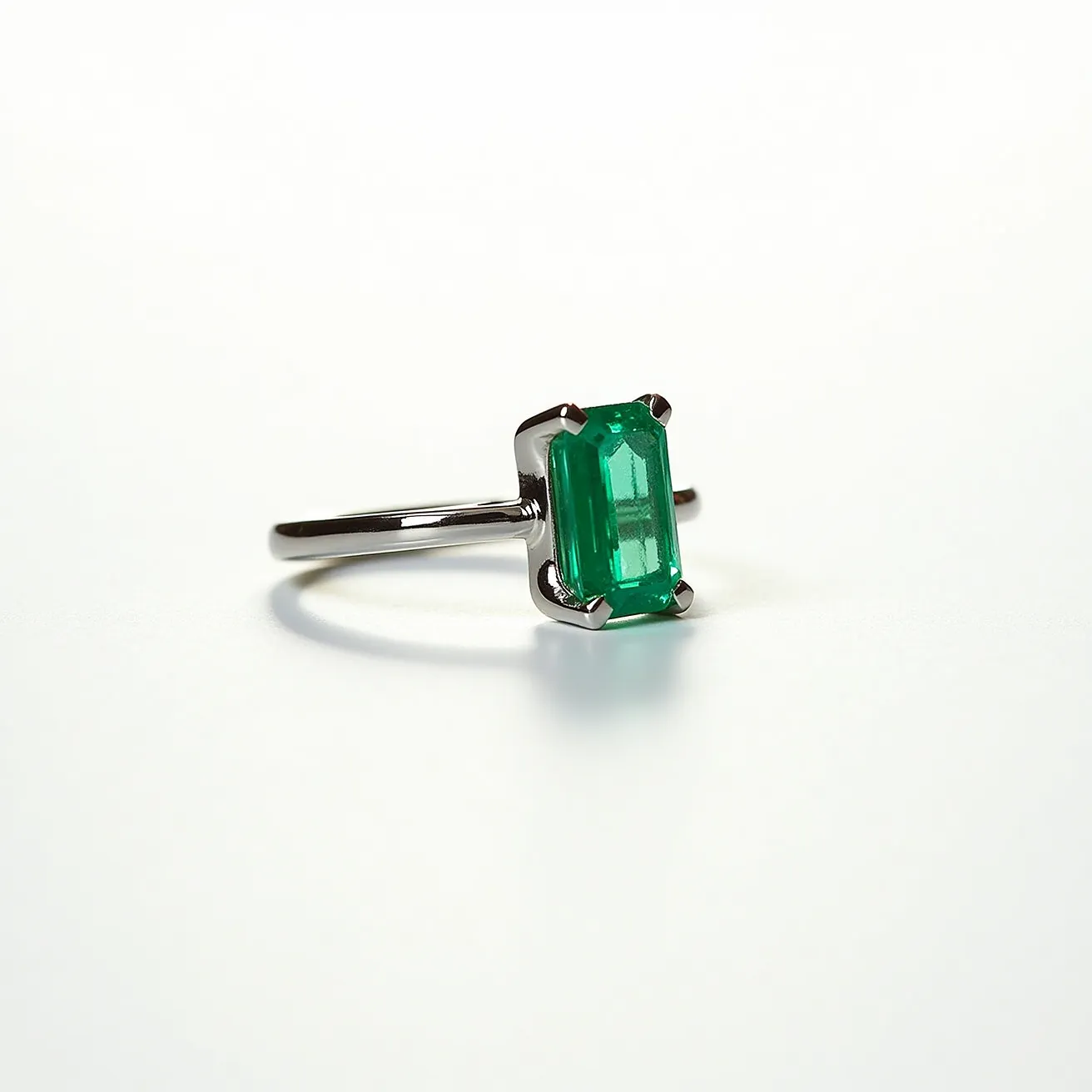 This engagement ring features a sleek, polished metal band, likely made of white gold or platinum, providing a sophisticated and modern appearance. At its center is a striking, emerald-cut green gemstone, which is securely held in place by a four-prong setting. The prongs enhance the stone's visibility while offering stability and security. The simple yet elegant design emphasizes the vibrant color and unique cut of the gemstone, making it the focal point of the ring.