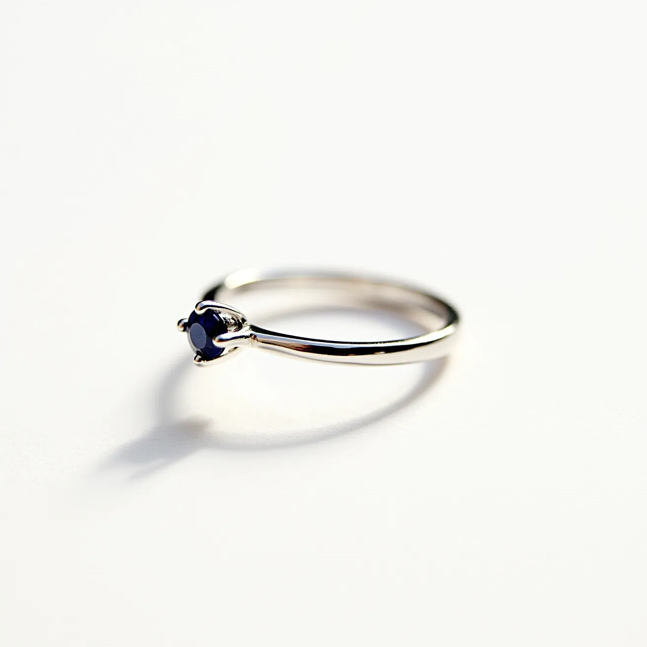 This engagement ring features a sleek, polished metal band, likely made of platinum or white gold, which provides a modern and elegant look. It is set with a deep blue gemstone, potentially a sapphire, expertly cut in a round shape to enhance its brilliance. The stone is secured in a classic four-prong setting that elevates it, allowing light to pass through and accentuate its vibrant color. The simplicity of the band highlights the stone as the focal point, creating a timeless and refined piece of jewelry.