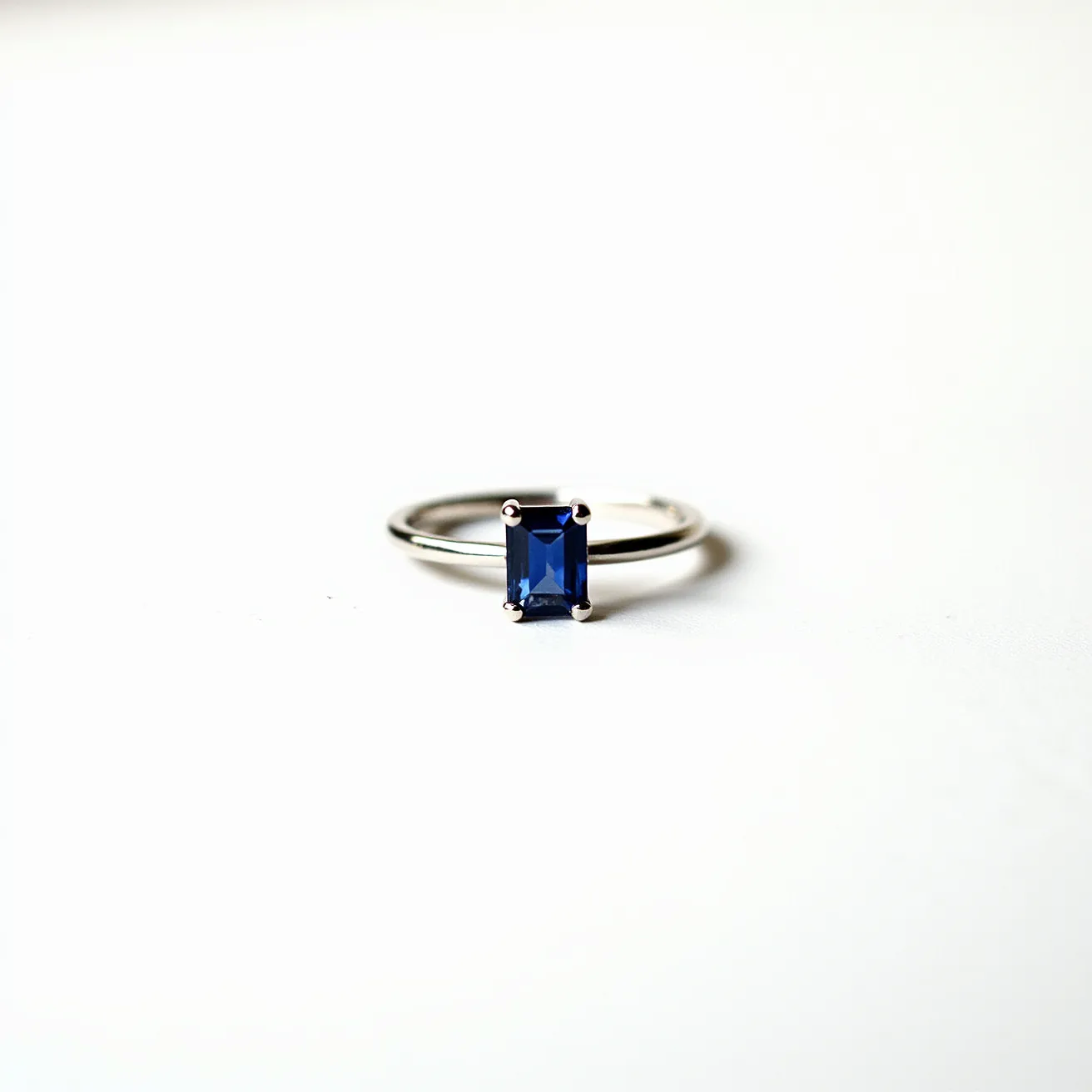 This engagement ring features a sleek metal band, likely crafted from white gold or platinum, which adds a modern and elegant touch. At its center is an exquisite rectangular-cut blue sapphire, securely held in place by a classic four-prong setting that accentuates the gem's vibrant hue and precise cut. The prongs not only ensure the sapphire's stability but also allow maximum light to enter the gem, enhancing its brilliance and sparkle. The minimalist design and quality materials make this ring a timeless symbol of commitment.