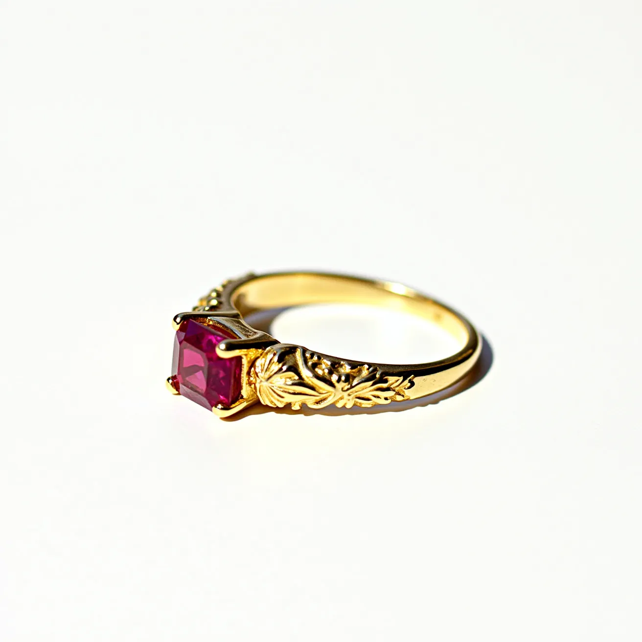 This engraved ring features a richly ornamented gold band with intricate leaf motifs adorning its sides. At its center, the ring holds a square-cut ruby gemstone set in a prong setting, which securely fastens the gem while allowing light to enhance its vibrant red hue. The gold band complements the deep color of the ruby, creating a striking contrast that highlights the gemstone's brilliance. The overall craftsmanship suggests elegance and attention to detail, making it a captivating piece of jewelry.