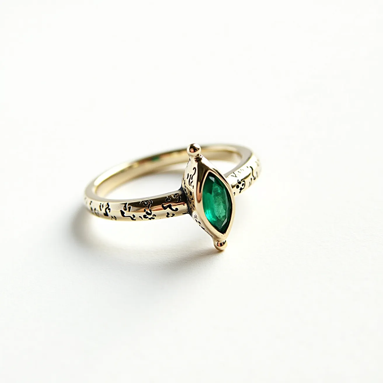 This engraved ring is crafted from a lustrous metal, likely gold, and features an intricate pattern along the band. Central to its design is a marquise-cut green gem, resembling an emerald, which is securely set in a bezel setting adorned with small prongs at the tips. The ring’s artistry is further accentuated by the delicate engravings, lending it an elegant and timeless appeal.