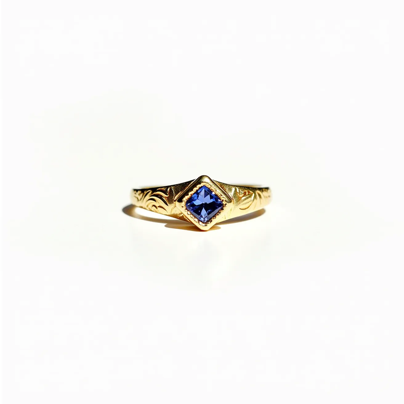 This engraved ring features a gold band adorned with intricate designs along its surface. At the center, it showcases a square-cut blue gemstone set in a bezel setting, adding a vibrant contrast to the gold. The ring embodies a classic elegance with its detailed craftsmanship and the rich hue of the gemstone, which forms a central focal point. The simplicity of the bezel setting enhances the beauty of the gem while providing a secure hold.