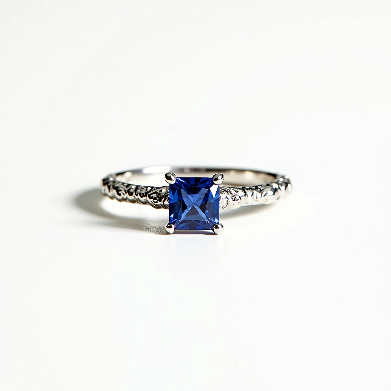 This engraved ring is crafted from a shiny metal, likely platinum or white gold, and features a detailed band with intricate engravings. At its center, the ring showcases a captivating square-cut blue gemstone, resembling a sapphire, securely held in place by a classic four-prong setting. The ring's design highlights both elegance and precision, with the gemstone serving as a stunning focal point. There are no additional attachments or clasps on the ring, allowing the stone and engravings to take center stage.