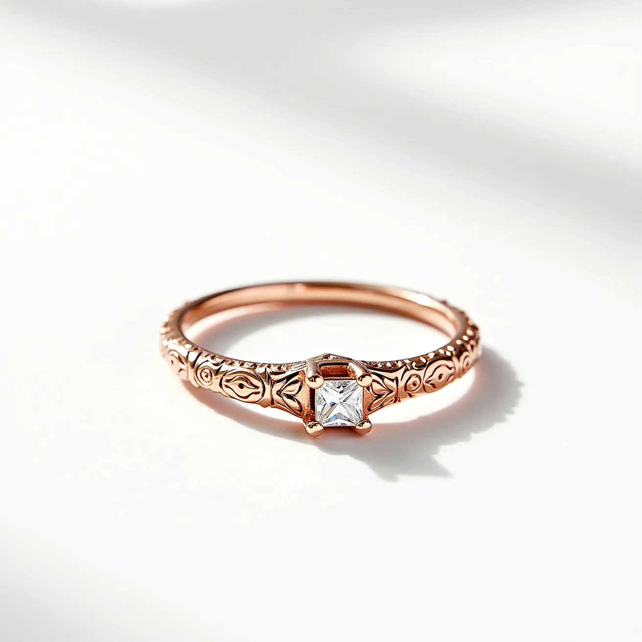 This engraved ring is crafted from a warm-toned metal, likely rose gold, and features intricate engravings along the band that add a touch of detailed elegance. At the center, a prominent square-cut gemstone, possibly a diamond, is set in a secure prong setting, showcasing its brilliance. The stone is elevated slightly above the band, emphasizing its prominence. The band itself is smooth and slender, providing a balanced contrast to the decorated area around the stone, embodying a blend of classic and ornate styles without any additional clasps or attachments.