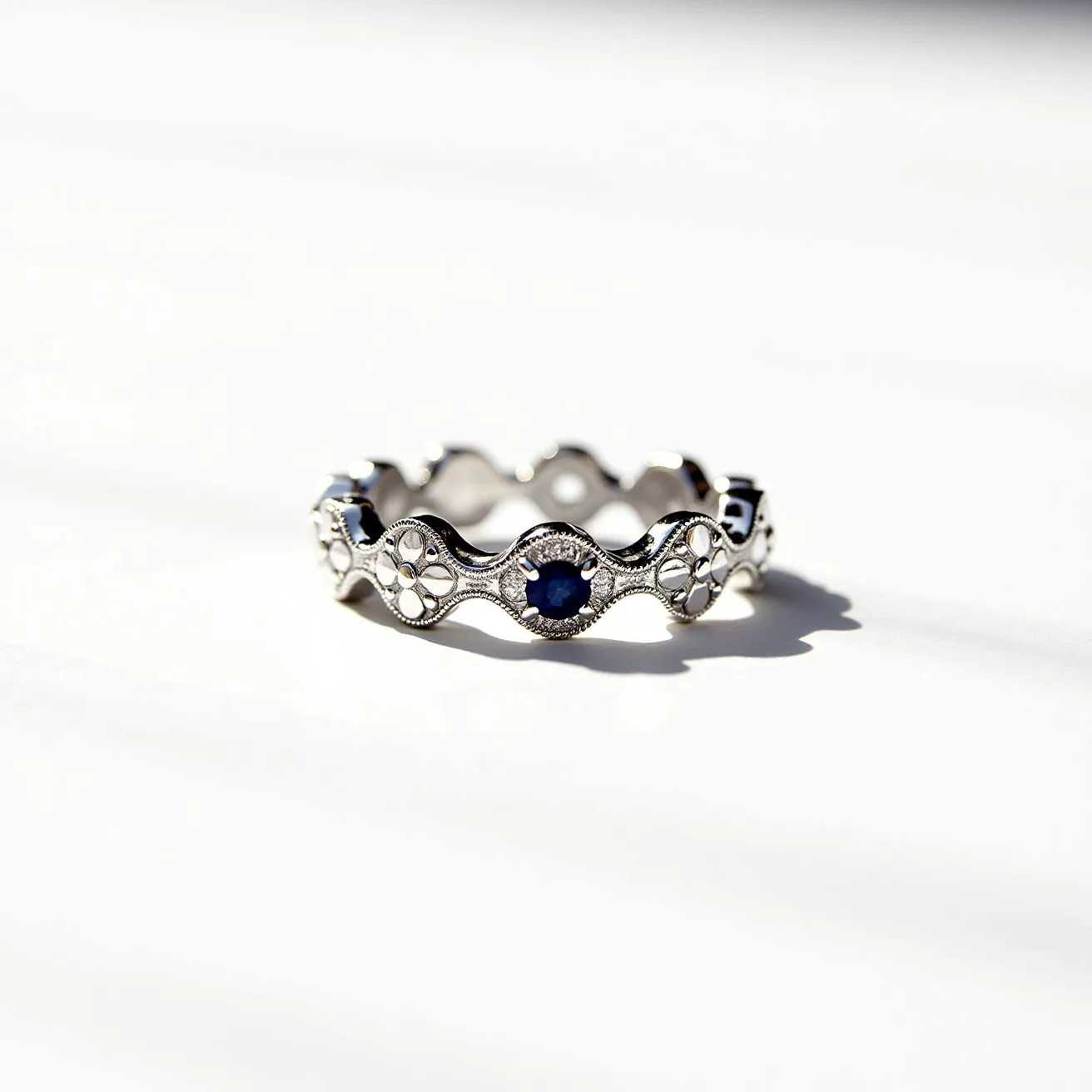 This eternity band ring features a sterling silver band with a wave-like design, intricately decorated with alternating round blue sapphires and floral motifs. The sapphires are set within round, milgrain-detailed bezel settings, adding a vintage touch. The continuous pattern exemplifies a classic eternity ring style, symbolizing endless love and commitment.