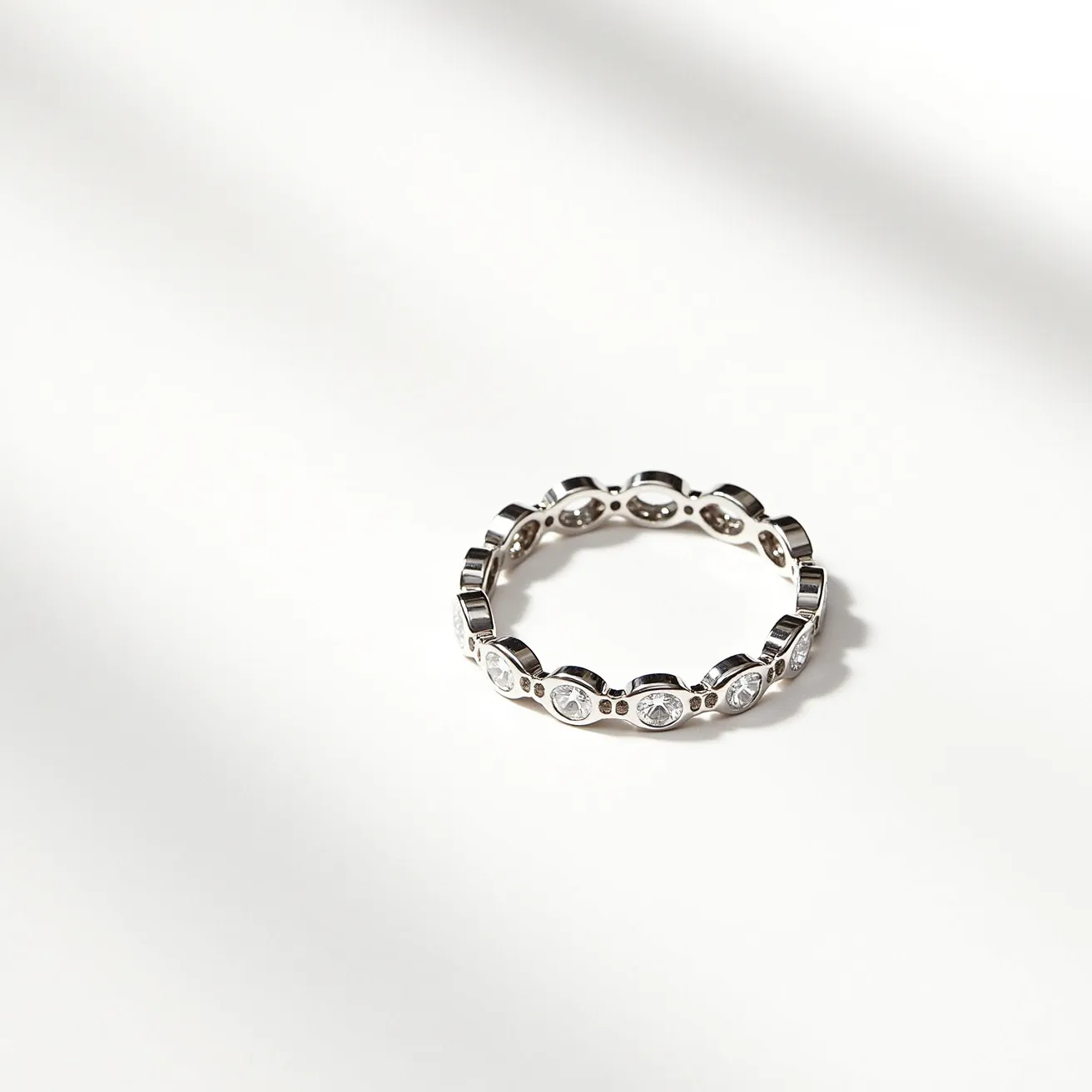 This eternity band ring features a continuous circle of clear, round-cut gemstones, likely diamonds or cubic zirconia, each set in individual bezel settings that provide both security and a sleek, modern appearance. The band itself is crafted from a reflective metal, possibly white gold or platinum, which enhances the brilliance of the stones. The design is seamless, indicative of the typical style of eternity bands, and there is no visible clasp or break in the continuity of the setting, emphasizing the symbolism of eternal love and commitment.