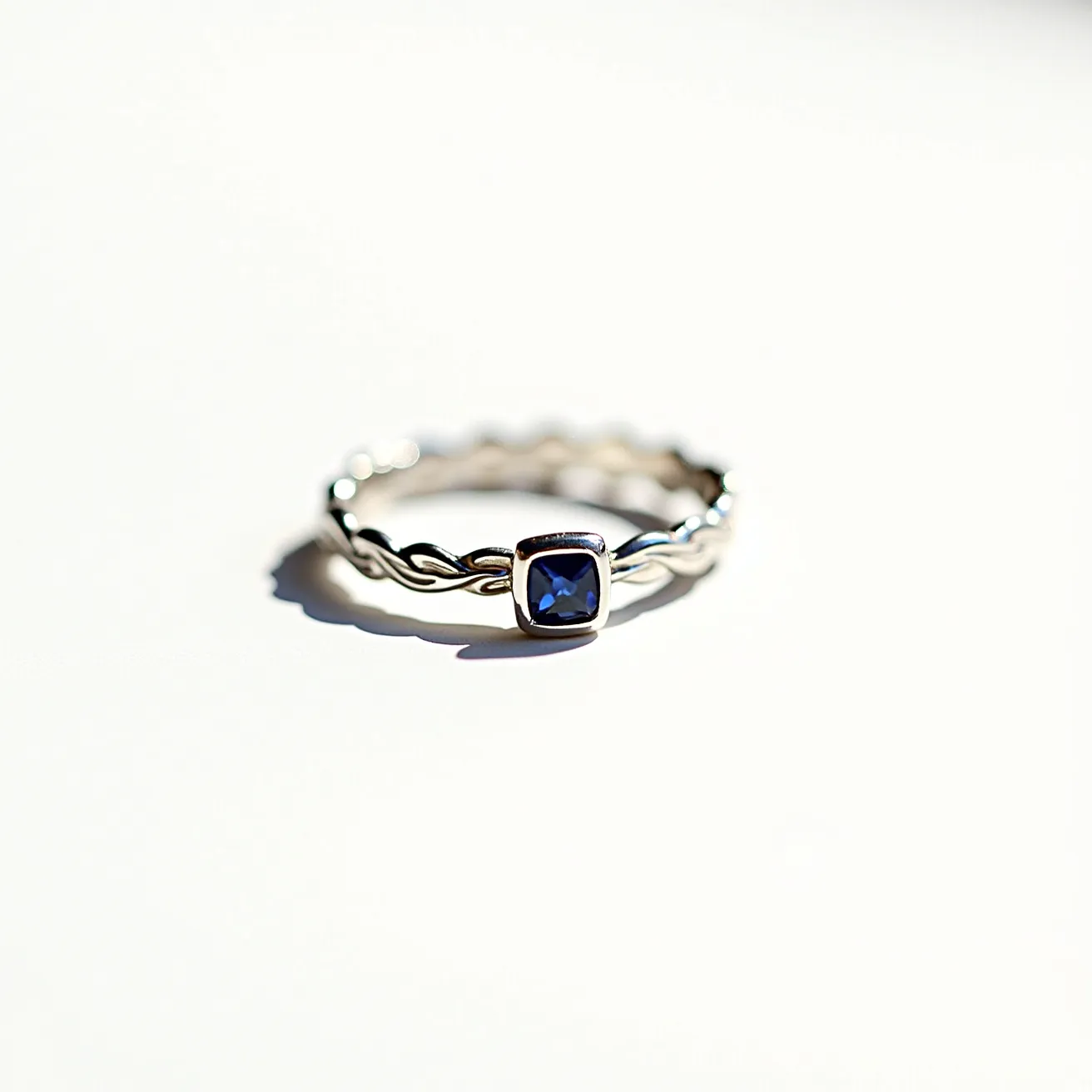 This eternity band ring features a braided metal band, likely crafted from silver or white gold, providing a sleek and elegant appearance. At its center is a prominent square-cut blue sapphire, which is bezel-set, enhancing the stone's sophistication and security while adding a vibrant splash of color to the design. The ring showcases a timeless style, with the intertwined band offering a delicate and refined touch, perfect for daily wear or as a statement piece. The sapphire's deep hue contrasts beautifully with the polished metal, creating a harmonious and attractive look.