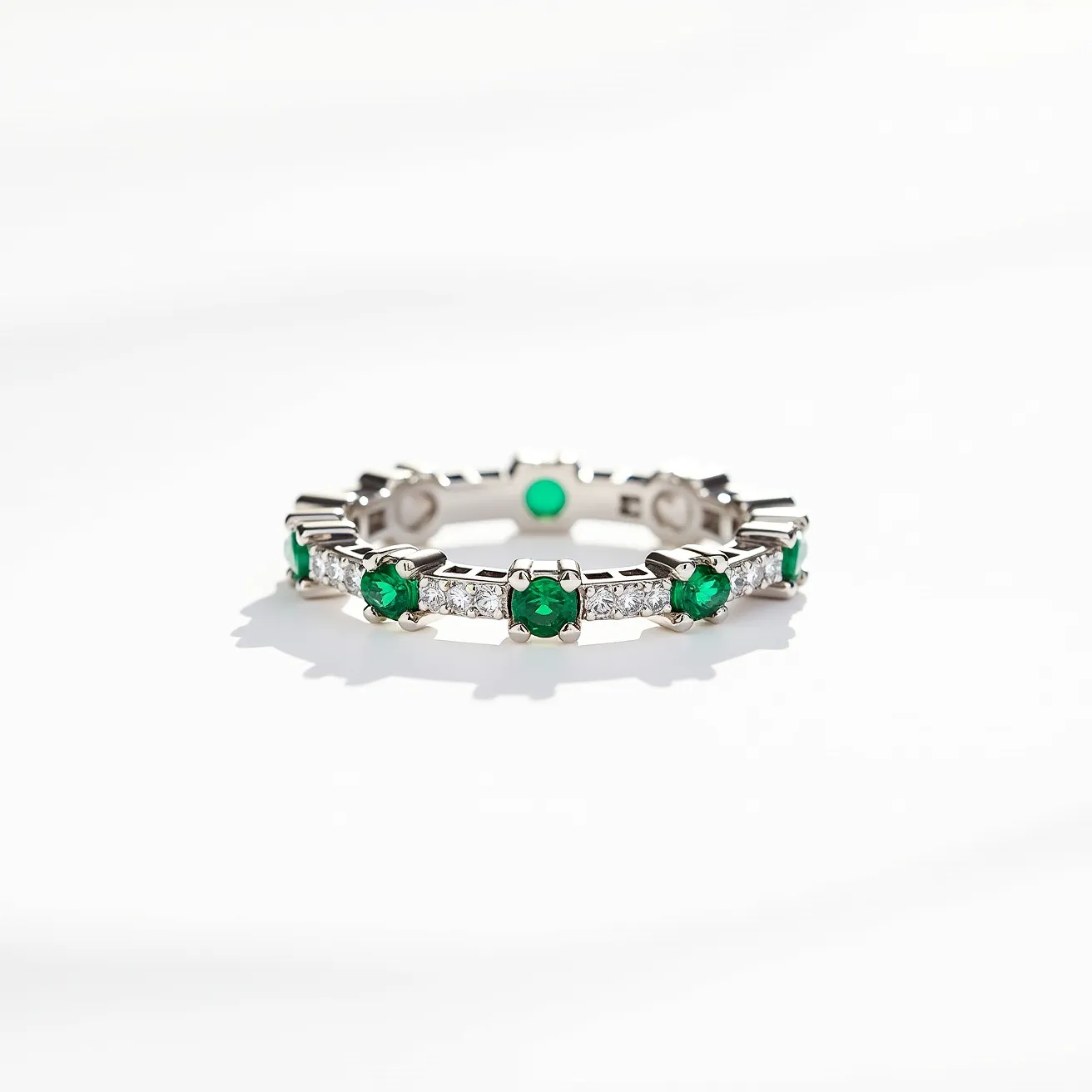 This eternity ring features a design crafted from a polished metal, likely white gold or platinum, with alternating round-cut green emeralds and round-cut white diamonds encircling the band. The emeralds are securely set in prongs, showcasing their vibrant green hue, while the diamonds between them add a brilliant sparkle to the overall appearance. The absence of a clasp signifies the continuous, unbroken nature of the eternity ring. The combination of materials and the precise setting enhances both the durability and elegance of the piece.