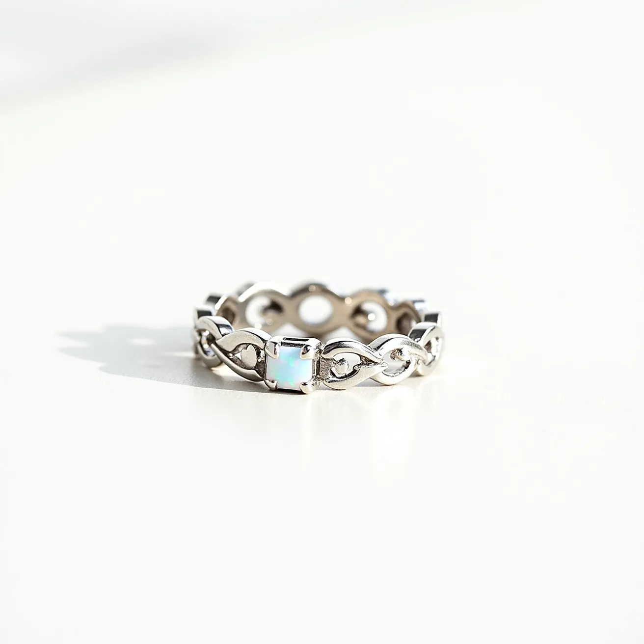 This eternity ring features a continuous band crafted from polished silver, showcasing an elegant looped design. The ring is adorned with a series of square-cut opals, each set within a secure bezel setting. The opals possess a subtle play of color, adding a touch of iridescence to the overall design. The interconnected pattern of the band enhances the flow of the piece, seamlessly integrating each gemstone without the need for a clasp or attachment.