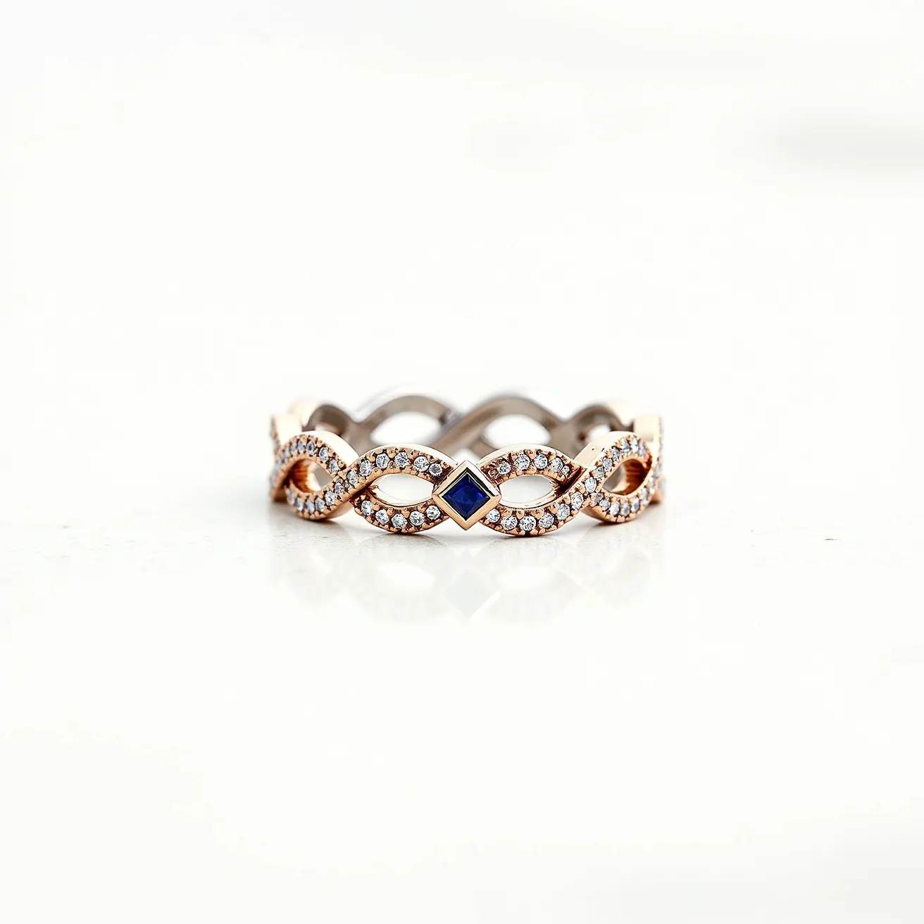 This eternity ring showcases an intricate design with intertwined bands crafted from a combination of rose gold and possibly white gold or platinum. It is beautifully adorned with small round-cut diamonds, each meticulously set in a pavé setting that enhances the continuous loop pattern. At the center of each loop is a square-cut blue sapphire, adding a striking contrast and a touch of elegance. The absence of a clasp signifies its seamless, continuous design, typical of eternity rings, which symbolizes everlasting commitment.