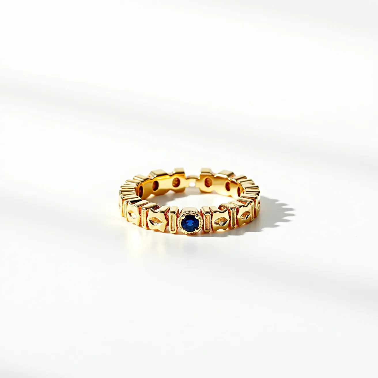 This eternity ring showcases a series of geometric patterns crafted in a lustrous gold material, which forms the band. At regular intervals around the band, there are striking blue sapphires set in bezel settings, adding vibrant color and elegance. The design is seamless and continuous, characteristic of an eternity band, symbolizing everlasting love and devotion.