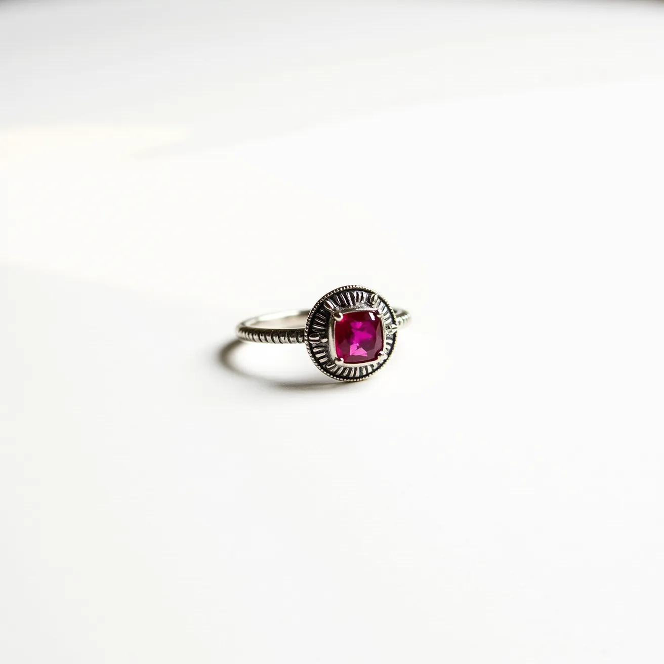 This fidget ring features a silver band with a detailed textured design surrounding a vibrant pink gem. The gem is square-cut and securely set in a prong setting, allowing for both stability and visibility of the stone. The silver band, possibly made of sterling silver, is intricately crafted to provide a tactile sensation, characteristic of fidget rings. The simplicity of its design, coupled with the striking central gemstone, makes it both a functional and aesthetically pleasing piece.