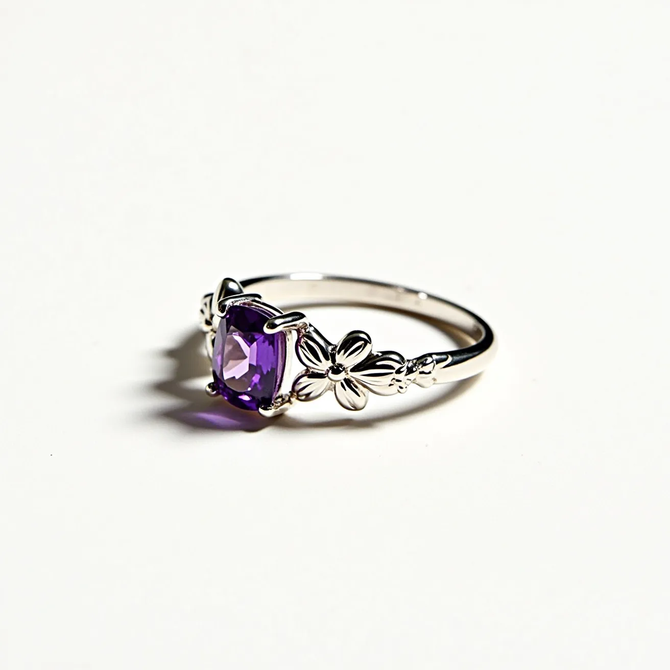 This floral ring features a stunning cushion-cut purple gemstone as its centerpiece, securely held by an elegant four-prong setting. The band appears to be crafted from a silver-toned metal, possibly sterling silver or white gold, enhancing its sophisticated look. Adorning the band are delicate floral motifs on either side of the gemstone, intricately detailed to add a touch of nature-inspired elegance without the need for additional clasps or attachments. The combination of the vibrant purple stone with the floral design creates an exquisite piece that balances timeless beauty with modern charm.