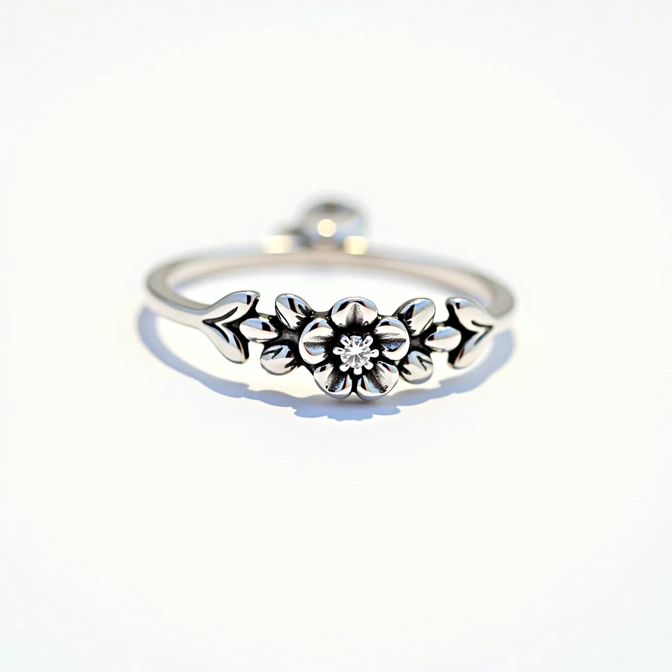 This floral ring features a delicate design with a prominent flower motif at its center. The ring appears to be crafted from a polished silver metal, giving it a sleek and shiny finish. The center of the flower houses a small, round-cut gemstone, likely a diamond, which is set in a prong setting to enhance its sparkle. The ring band itself is simple and elegant, complementing the intricacies of the floral design without overshadowing it. The detailing on the petals and leaves adds a touch of finesse, making the piece both understated and charming.