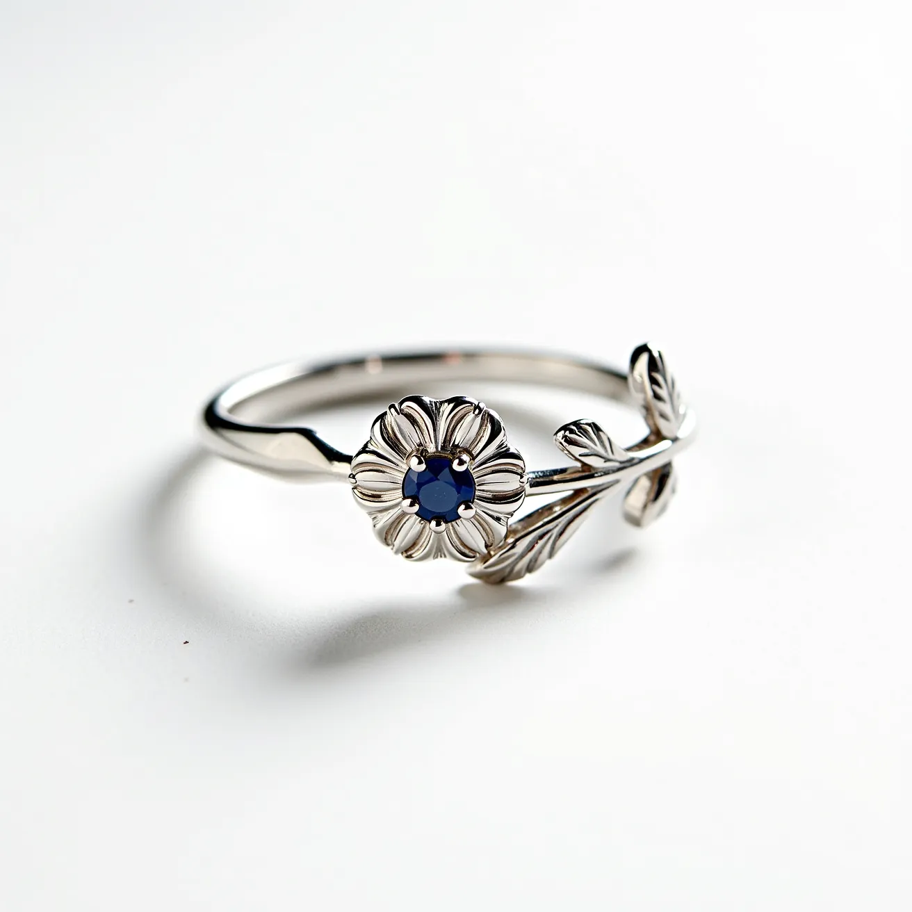 This floral ring features a delicately designed band resembling a vine or branch, crafted from a polished silver material. The focal point is a flower motif, which artistically incorporates a central blue gemstone. The gem appears to be a round cut and is elegantly set in a claw setting, emphasizing its vibrant hue. The ring's intricate detailing includes leaves along the band, complementing the floral design. There is no visible clasp or additional attachment, with the design focusing on simplicity and elegance.