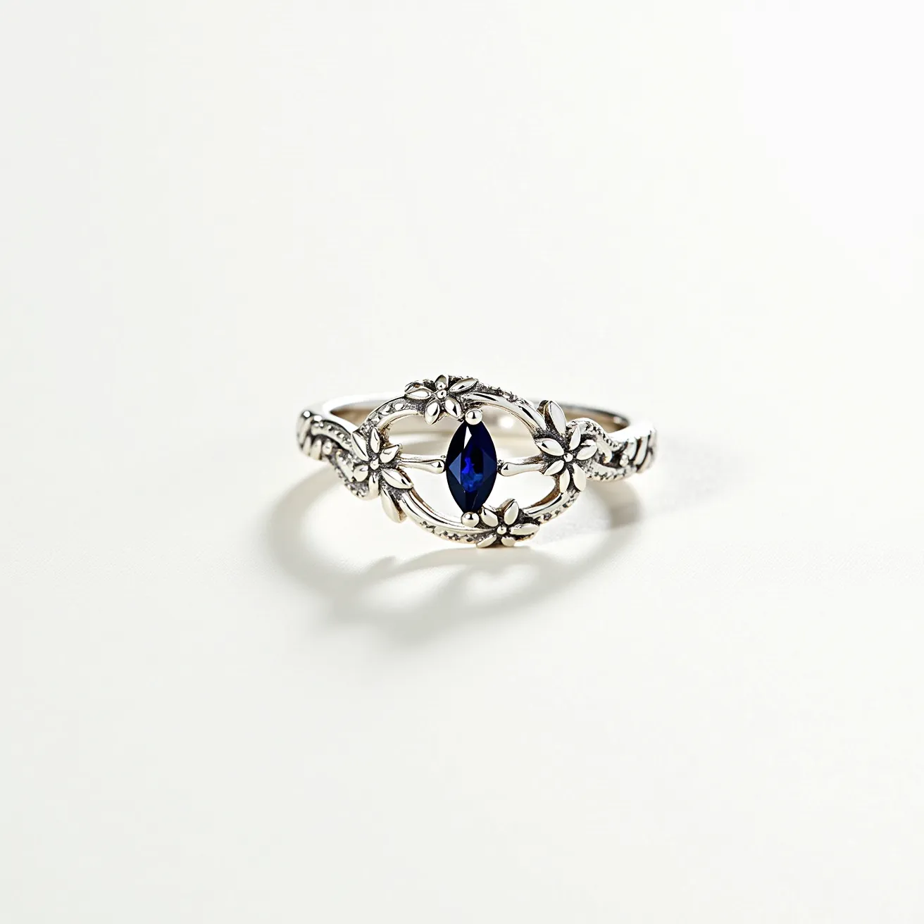 This floral ring features a delicate, intricately designed silver band adorned with small floral motifs circling a striking marquise-cut blue gemstone. The central stone is securely set within the band, with its vibrant hue creating a captivating contrast against the metallic backdrop. The floral embellishments extend along the band, enhancing the piece’s elegant and nature-inspired aesthetic. The unified design seamlessly integrates the gem into the ring, showcasing a refined and harmonious appearance.