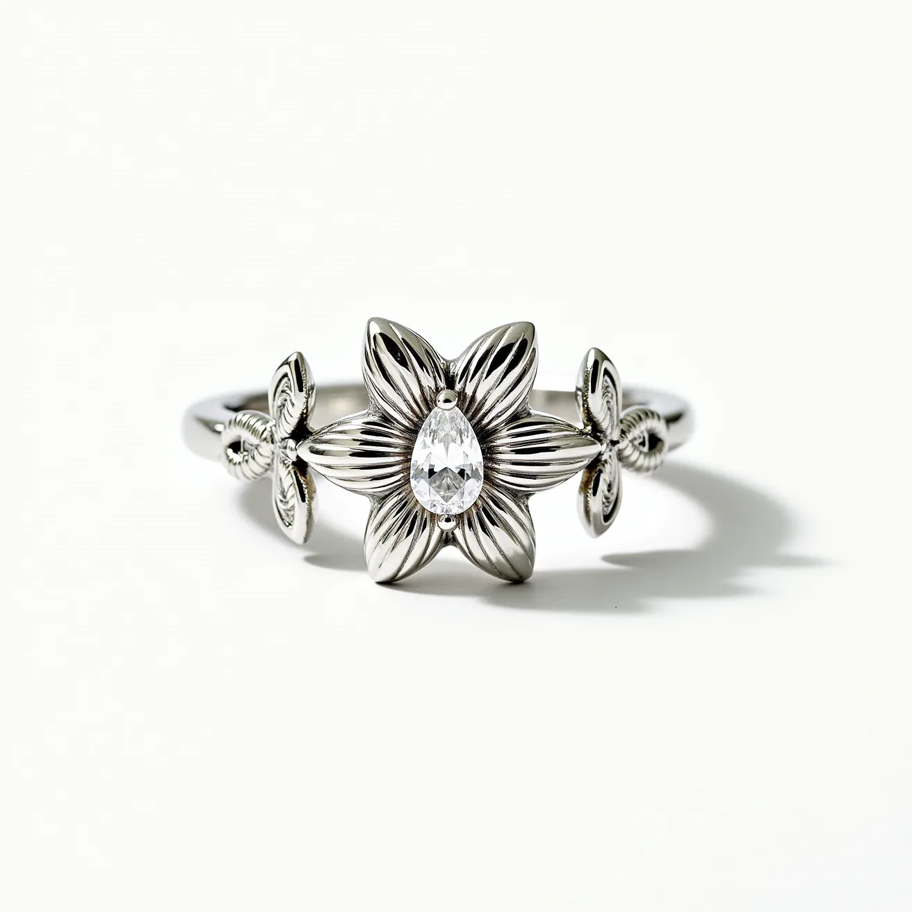 This floral ring features a striking central flower design with a polished finish, crafted from a silver-hued metal. At the heart of the flower is a pear-shaped gemstone, likely a diamond or similar clear stone, set in a delicate prong setting that accentuates its brilliance. Flanking the central bloom are two smaller floral motifs, adding to the ring's intricate and harmonious design. The band is smooth and seamlessly integrated with the floral elements, offering a graceful and fluid appearance. The overall composition of the ring highlights elegance and detailed craftsmanship.