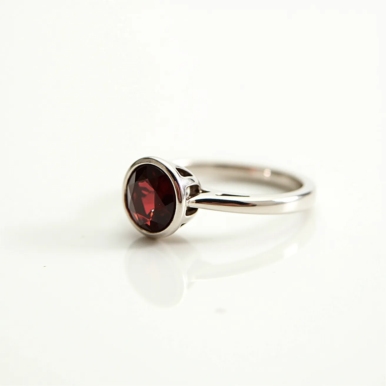 This garnet engagement ring features a striking, deep red garnet gemstone set in a bezel setting, which securely encases the stone. The gemstone is cut in a round shape, enhancing its rich color and brilliance. The band is crafted from a polished metal, likely platinum or white gold, complementing the color and style of the garnet. Its smooth, sleek design and secure gemstone setting make it both elegant and timeless in appearance.