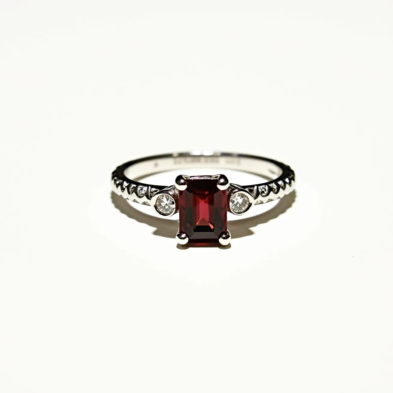 This garnet engagement ring features a striking rectangular cut garnet as the centerpiece, securely held in place by four prongs. The deep red hue of the garnet contrasts beautifully with the band, which is likely made of a silver-tone metal, enhancing its elegance. Flanking the center stone on either side are small round clear gemstones, possibly diamonds or diamond simulants, set in bezel settings that add a touch of sparkle. The band is adorned with a series of small, round clear stones set within a delicate channel setting, creating a continuous line of brilliance along the surface. This combination of materials and settings results in a sophisticated and timeless design.