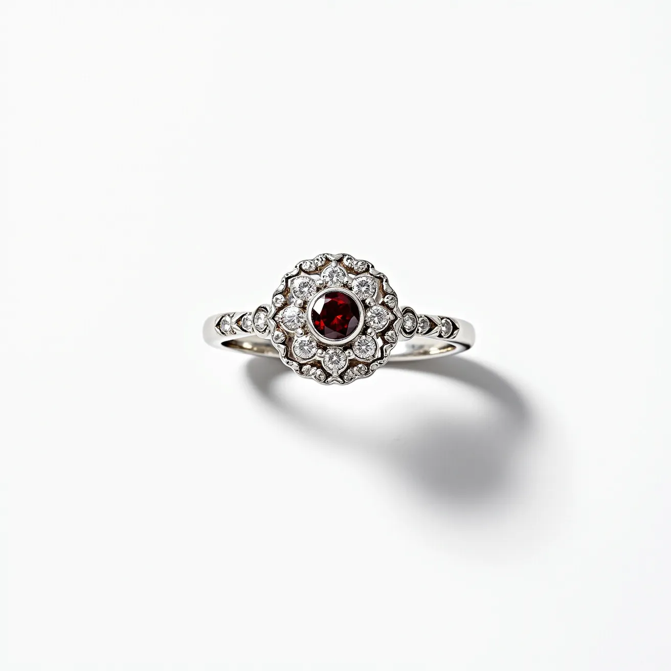 This garnet engagement ring features a central round-cut garnet, set in a bezel style. Surrounding the garnet is an ornate halo of round, clear diamonds, each held in place by prong settings, which enhance the ring's overall sparkle and elegance. The band is made of a shiny silver metal, likely white gold or platinum, and includes additional diamond accents along the shoulders. This design combines vintage charm with modern sophistication, creating a captivating and timeless piece.