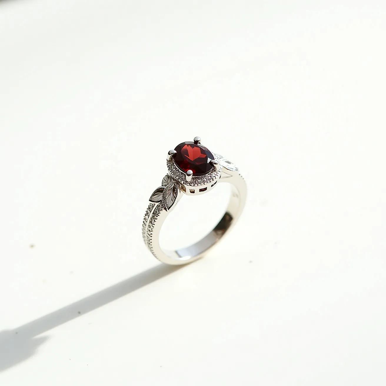 This garnet engagement ring features a striking oval-cut garnet gemstone, set prominently at the center. The garnet is surrounded by a halo of small, sparkling diamonds, accentuating its rich, red hue. The band is made of white gold, intricately designed with leaf motifs and detailed milgrain edging, adding an elegant touch to the overall appearance. The setting is a classic prong style, securing the garnet and ensuring its prominence. The ring combines the boldness of the garnet with the subtlety of the white gold and diamond accents, creating a harmonious and sophisticated piece.