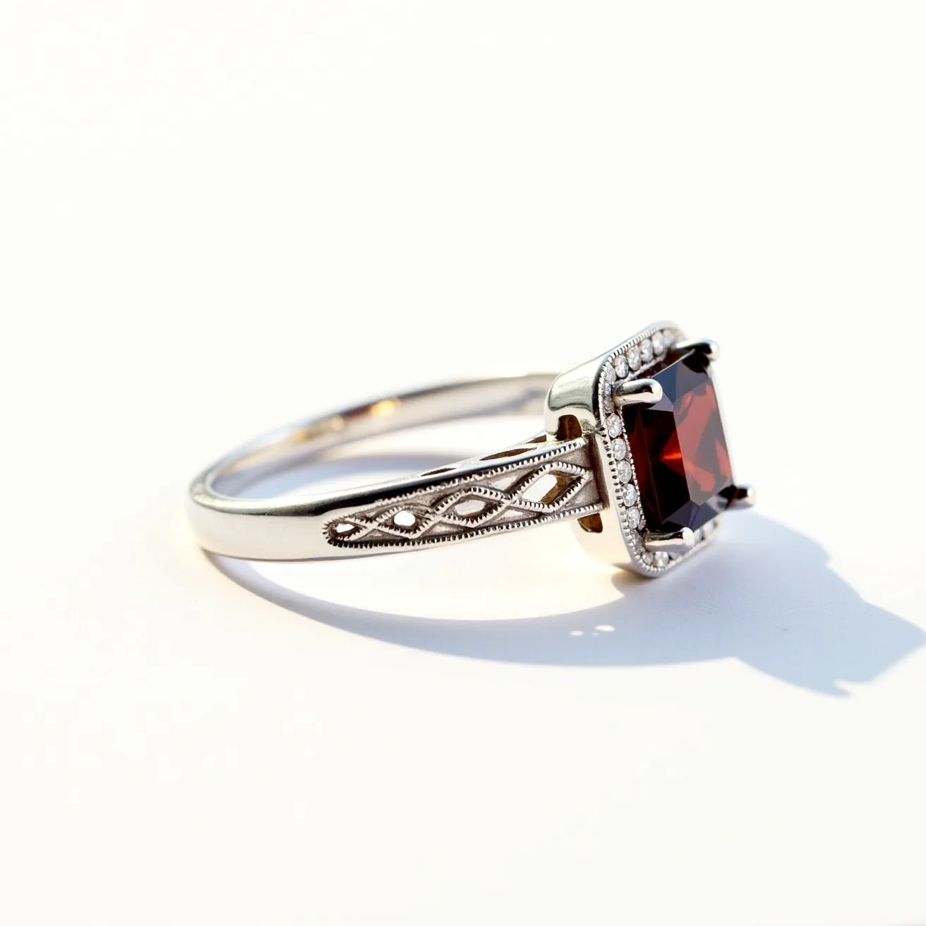 This garnet ring features a striking central square-cut garnet, set in an elegant prong setting. The band is crafted from a lustrous metal, possibly silver or white gold, accentuated with intricate filigree designs that run along the sides. These ornate details enhance the overall aesthetic, adding a touch of vintage charm. The setting is complemented by a row of small, sparkling stones encircling the garnet, likely diamonds or cubic zirconias, providing a brilliant contrast to the deep red hue of the garnet. The combination of the central gemstone and decorative elements creates a visually captivating piece.