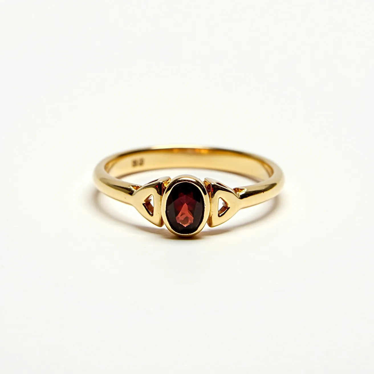 This garnet ring features a rich, oval-cut garnet set at its center, which is the focal point of the design. The garnet is secured within a gold setting, enhancing its deep red hue. The band is crafted from gold, offering a sleek and polished appearance that complements the stone. On either side of the garnet, there are decorative gold elements that add a touch of elegance to the overall design. The ring band appears to be smooth and continuous, suggesting a traditional ring structure without any visible clasps or attachments.