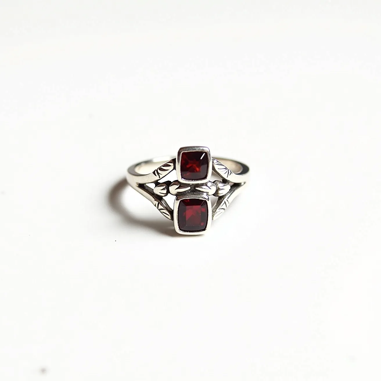 This garnet ring features two distinct square-cut garnet stones, each securely set in a bezel setting, which enhances their deep red color while providing a modern, streamlined look. The band is crafted from polished silver, displaying a delicate design with subtle leaf-like engravings that add an elegant touch to the piece. The dual stone arrangement is symmetrical, offering a balanced aesthetic that complements the intricate detailing on the band, showcasing both craftsmanship and style.