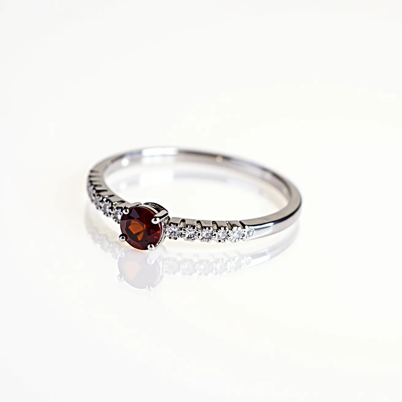 This garnet ring features a striking round-cut garnet gemstone as its centerpiece, set in a lustrous metal band that appears to be white gold or platinum. The garnet, with its deep red hue, is securely held in place by four prongs, enhancing its brilliance and visibility. Adorning the band on either side of the central garnet are several small, round-cut white diamonds, also prong-set, which add a touch of elegance and sparkle. The band is sleek and smooth, designed to highlight the gems without distraction. The combination of the rich garnet and sparkling diamonds creates a timeless and sophisticated look.