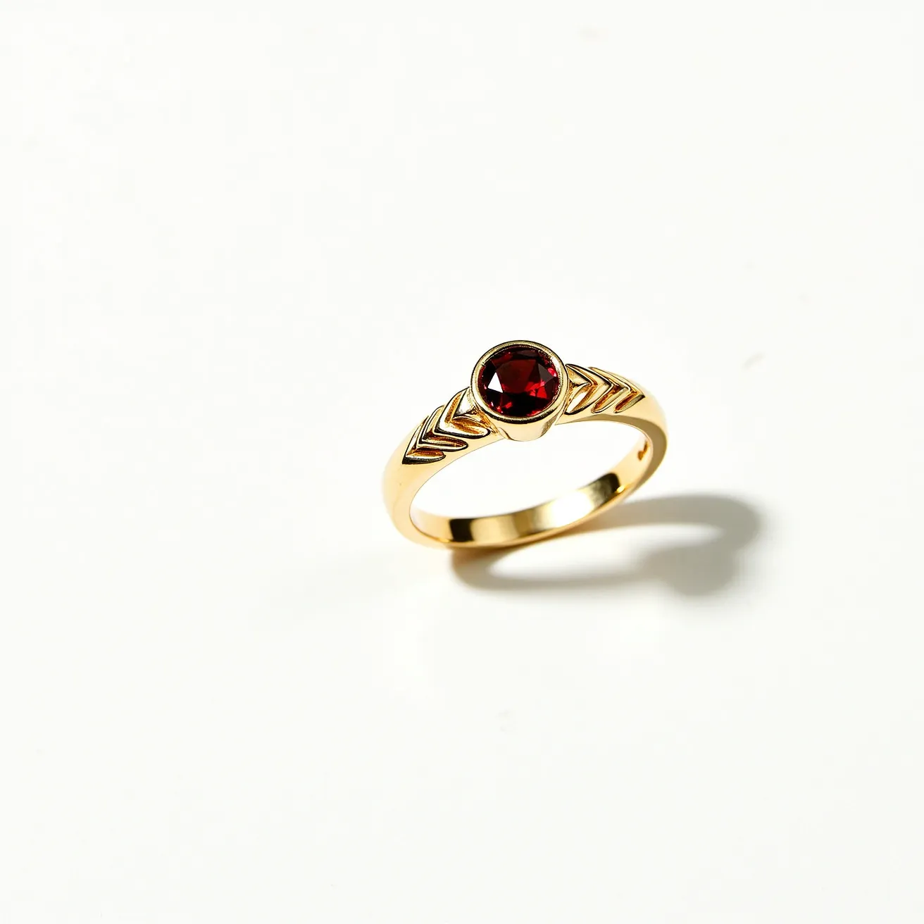 This garnet ring features a striking round-cut garnet set into a gold band, highlighting the stone's deep red hue. The stone is securely held in a bezel setting, which enhances its simplistic elegance and contemporary style. The gold band is adorned with a symmetrical arrow-like pattern on either side of the garnet, adding a touch of intricate design to the overall appearance. The choice of materials and geometric details give this ring a modern yet timeless quality, making it a standout accessory.