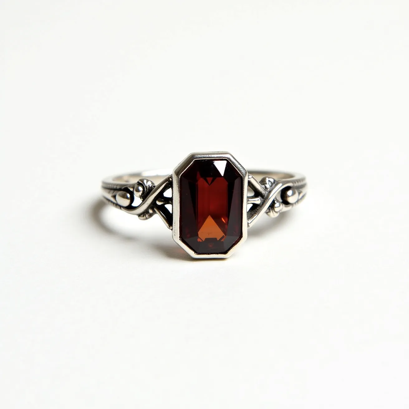 This garnet ring features a striking, rectangular emerald-cut garnet set in a polished metal band, likely made of silver due to its color and finish. The garnet's deep red hue is complemented by an ornate band design, which involves intricate scrollwork and beading on either side of the central stone, enhancing its vintage aesthetic. The garnet is held securely in a bezel setting that wraps around the stone's edges, providing both protection and an elegant frame. The ring's craftsmanship highlights the rich color of the garnet, making it a stunning piece of jewelry.