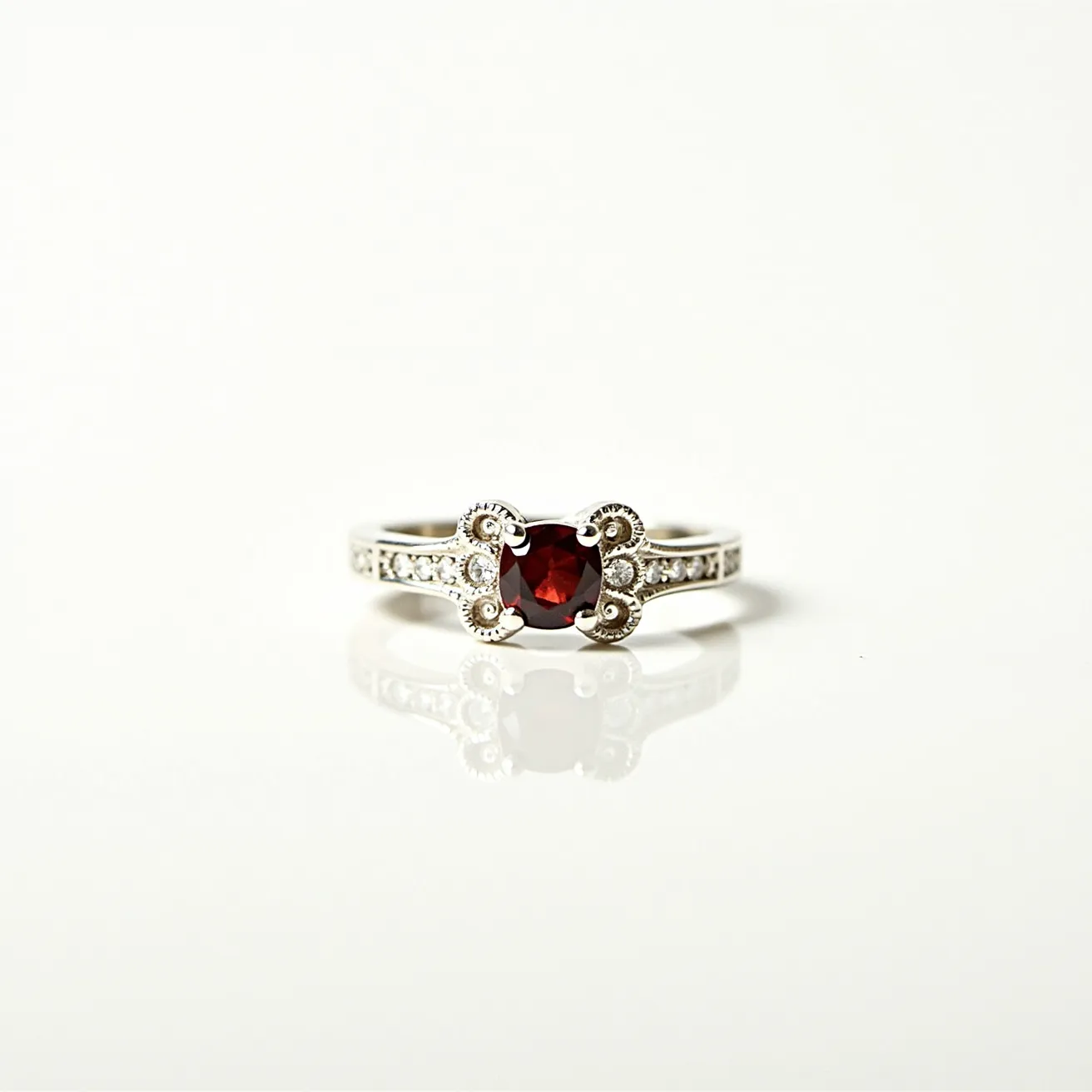 This garnet ring features a deep red garnet gemstone set in a cushion cut, which is elegantly secured by four prongs forming a distinctive scroll-like setting. The band is likely made of a silver or white gold material, complementing the rich hue of the garnet. It is adorned with small clear stones, possibly diamonds or diamond simulants, channel-set along the shoulders, adding a touch of sparkle and enhancing the ring’s elegance. The overall design combines vintage elements with a classic aesthetic, making it a timeless piece of jewelry.