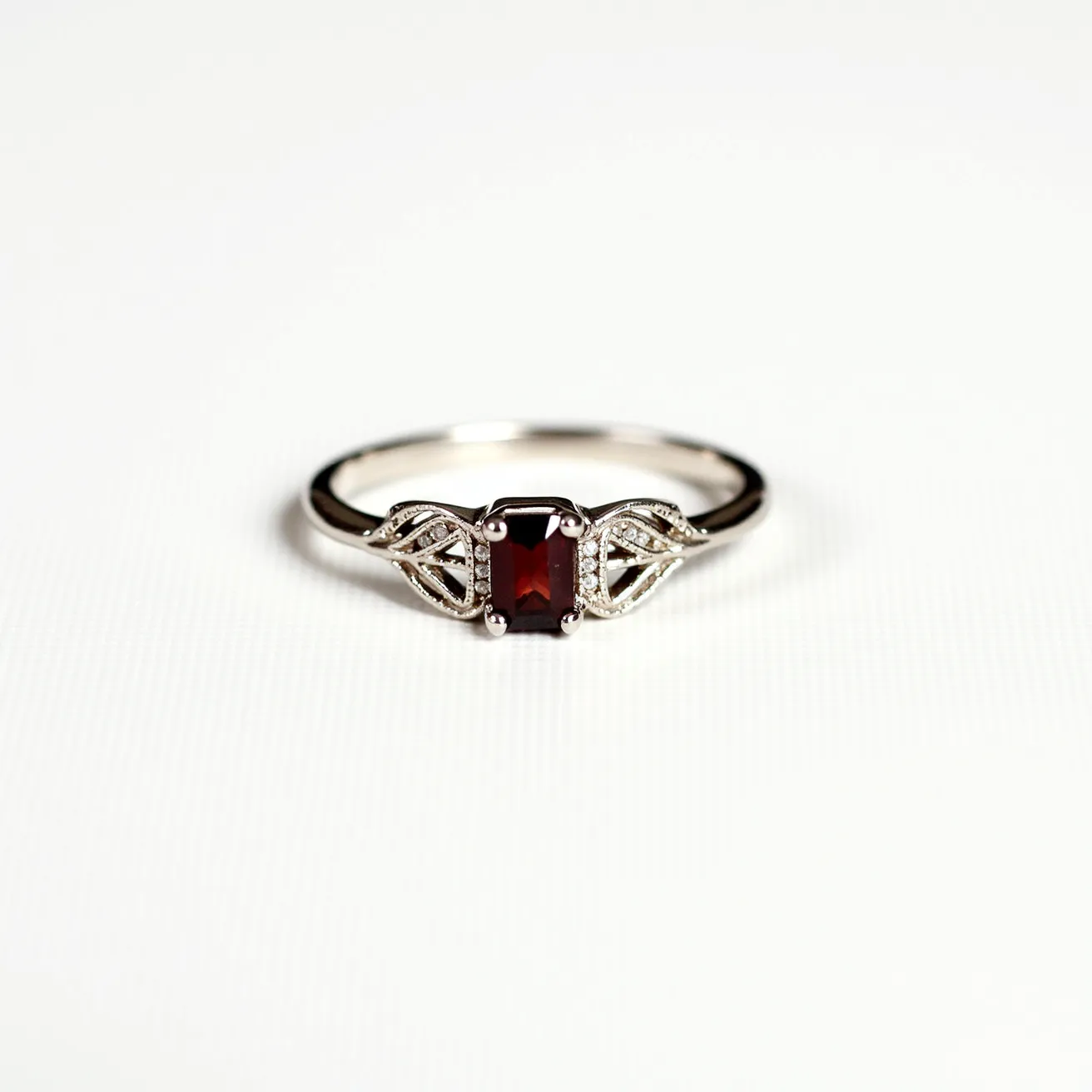 This garnet ring features a central rectangular-cut garnet gemstone with a deep red hue. The stone is set in a classic prong setting, which secures and showcases its vibrant color. Flanking the garnet are delicate, leaf-like designs crafted from the ring's metal, possibly white gold or platinum, and accented with small, round brilliant-cut diamonds that add a subtle sparkle. The band is smooth and polished, providing a sleek and elegant appearance. The design elements harmonize to create a refined and sophisticated piece of jewelry.