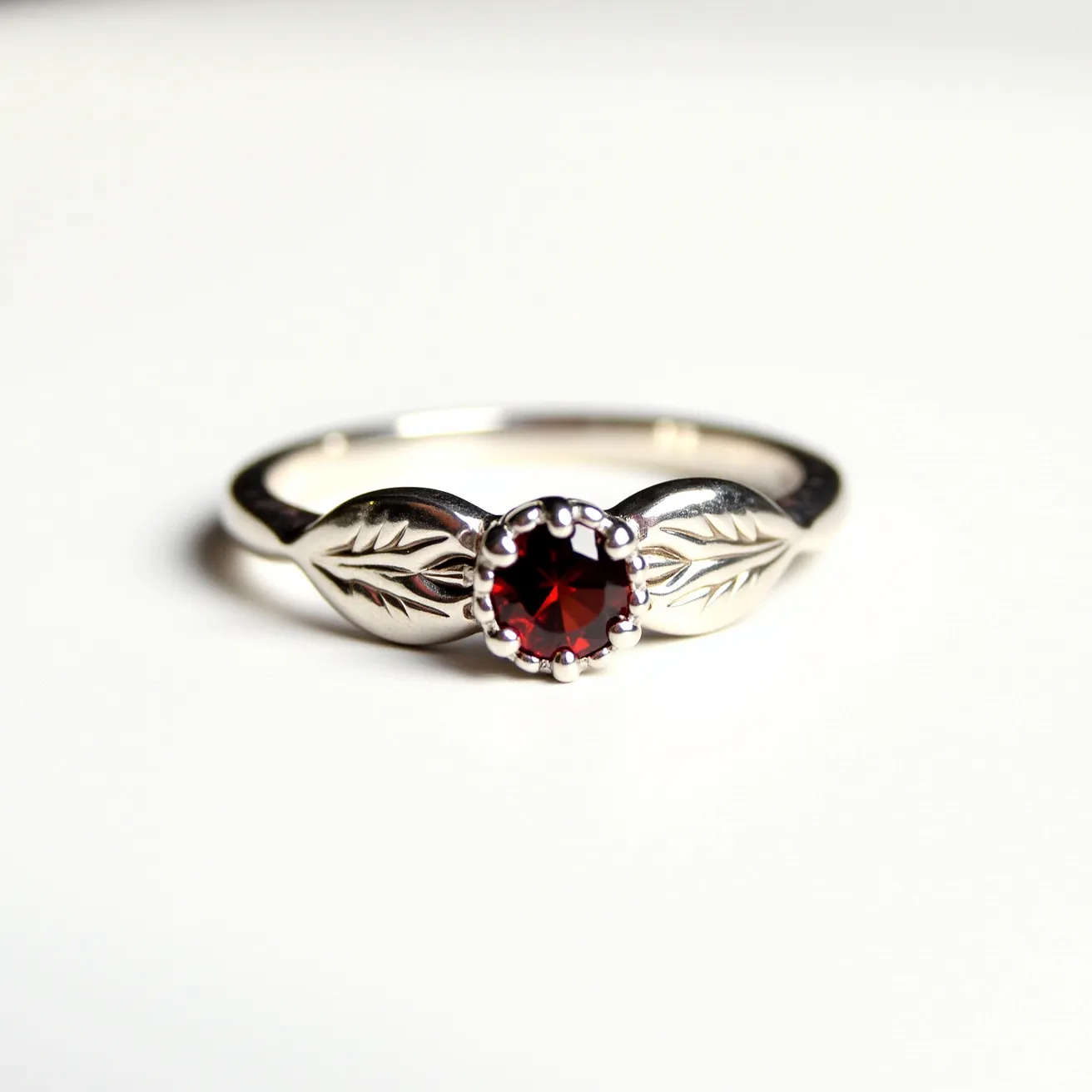 This garnet wedding ring features a beautifully faceted round garnet set in what appears to be a silver band. The garnet is held in place by a classic prong setting, which accentuates the stone's rich red hue. The band is embellished with detailed leaf motifs on either side of the central gemstone, adding an elegant and nature-inspired design element. The polished finish of the silver band complements the deep red garnet, creating a harmonious and timeless piece.