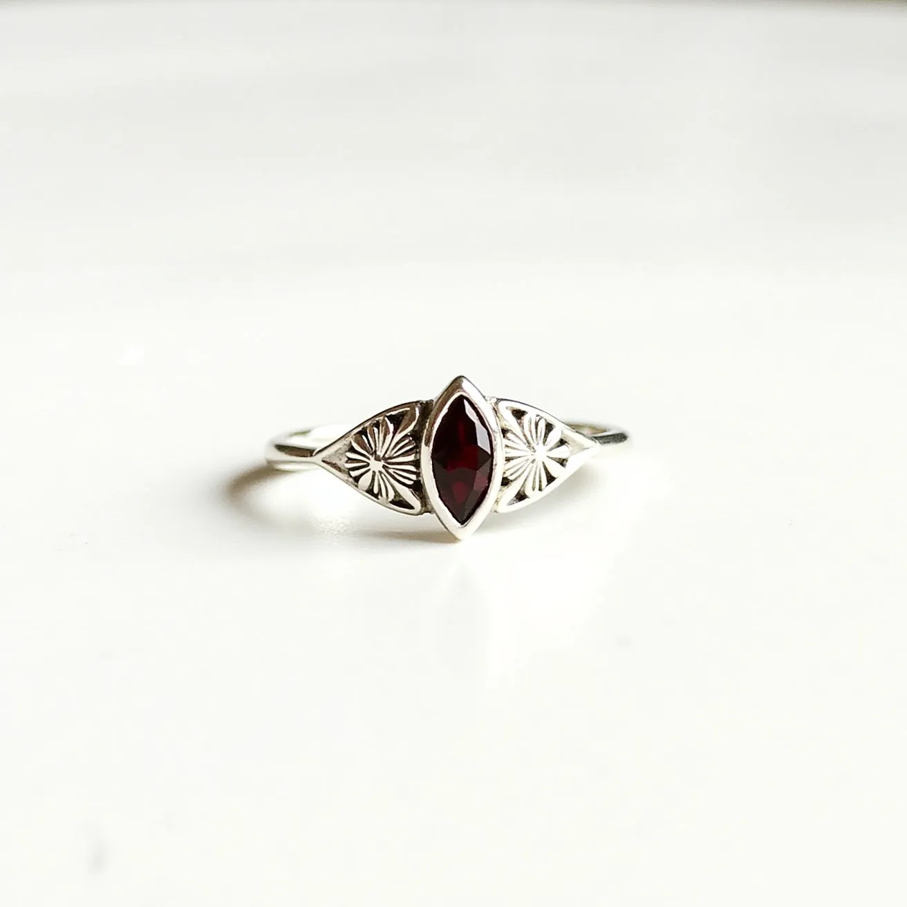 This garnet wedding ring features a marquise-cut garnet set at its center, elegantly framed in a bezel setting. The band is crafted from a silver-toned metal, showcasing an intricate design with engraved floral motifs on either side of the garnet. The delicate band complements the central stone, enhancing its deep red hue. There is no clasp or additional attachments, allowing the garnet and the unique design of the band to stand out distinctly.