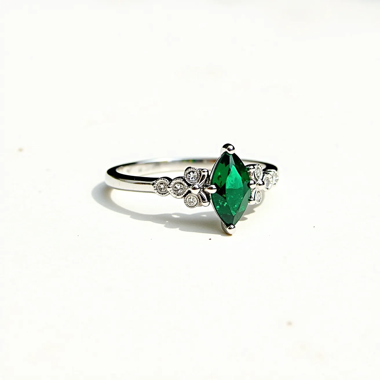 This gemstone engagement ring features a striking marquise-cut green gem as its central piece, set in a delicate prong setting. The band appears to be made of a polished white metal, likely white gold or platinum, which complements the vivid color of the centerpiece. Flanking the central stone are decorative floral motifs adorned with small, round-cut diamonds that add a touch of sparkle and elegance. The intricate arrangement of the gems and the refined craftsmanship reflect a classic yet unique design, perfect for marking an engagement with a sense of timeless beauty.