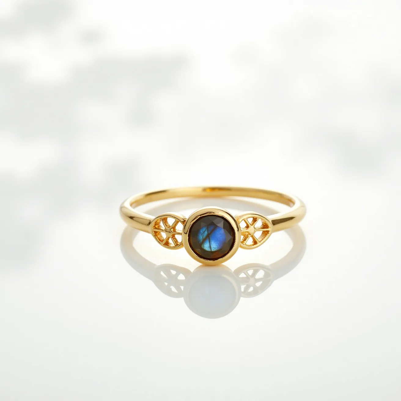 This gemstone ring features a round, blue labradorite gemstone set centrally in a bezel setting, highlighting its unique luminescent qualities. The band appears to be crafted from gold, possibly gold-plated or solid gold, providing a warm contrast to the cool hues of the labradorite. On either side of the gemstone, intricate openwork detailing complements its design, adding an elegant touch without overpowering the central stone. The simplicity and elegance of the ring make it suitable for both everyday wear and special occasions.