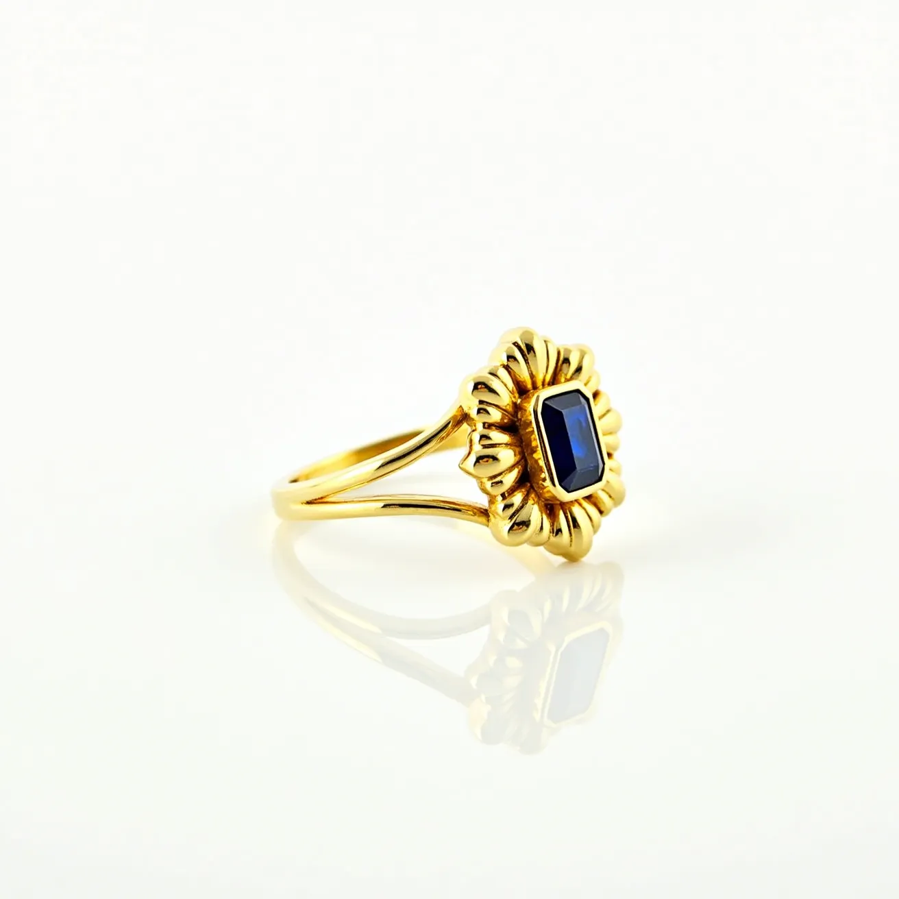 This gold ring features a delicately crafted band with an ornate design that highlights a dark blue rectangular stone, resembling a sapphire. The stone is set within a unique floral bezel setting, which adds an additional layer of elegance and complexity to the piece. The ring showcases a split shank design, contributing to its sophisticated and intricate appearance. The deep hue of the stone contrasts beautifully with the warm tones of the gold, making it a striking and exquisite piece of jewelry.