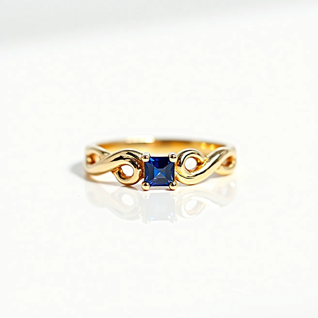 This gold thumb ring features a delicate design crafted from polished gold, showcasing a central square-cut blue gemstone, likely a sapphire, set in a four-prong setting. The band is elegantly twisted with flowing, openwork loops, adding a touch of intricate detail to its appearance. Overall, the combination of the gemstone’s deep blue hue with the gold’s warm tone creates a visually appealing and sophisticated piece.