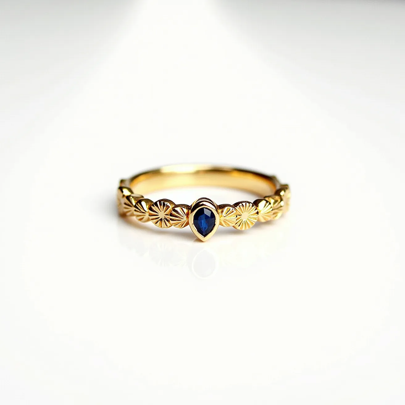 This gold wedding band is crafted with intricate designs that resemble floral patterns etched along its surface. It features a deep blue sapphire at its center, which is pear-shaped and set in a secure bezel setting. The band itself appears to be made of yellow gold, enhancing the elegant and classic appearance of the ring. There are no clasps or attachments visible, making it a seamless, continuous piece.